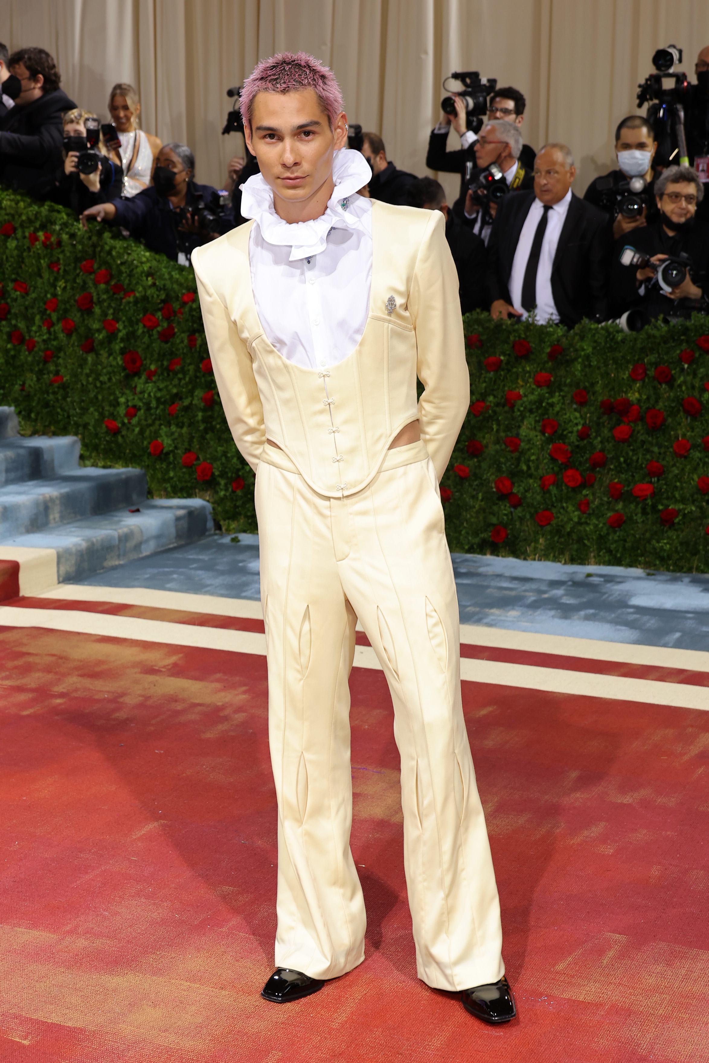 Person in a tailored, cream-colored suit with a high ruffled collar stands on a red carpet. Background features photographers and greenery