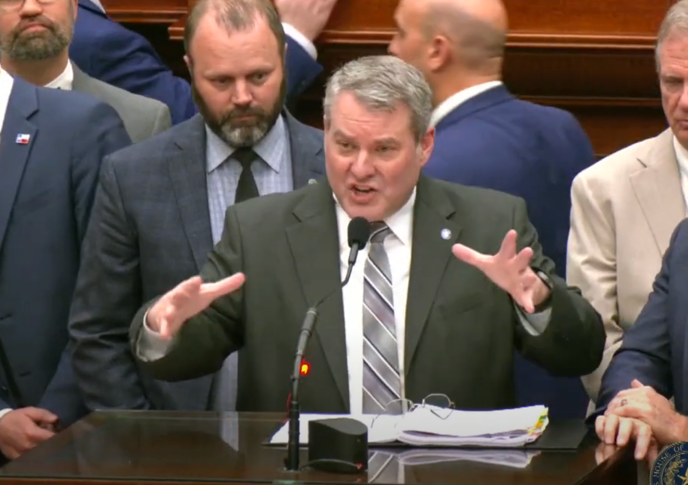 A man in a suit gestures while speaking at a podium in a formal setting, surrounded by other men in suits