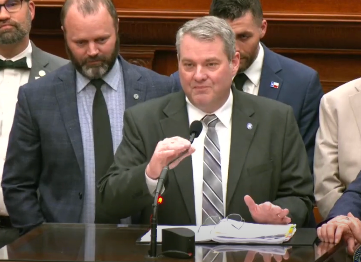 A man in a suit speaks at a podium surrounded by others, appearing to deliver a formal statement or address