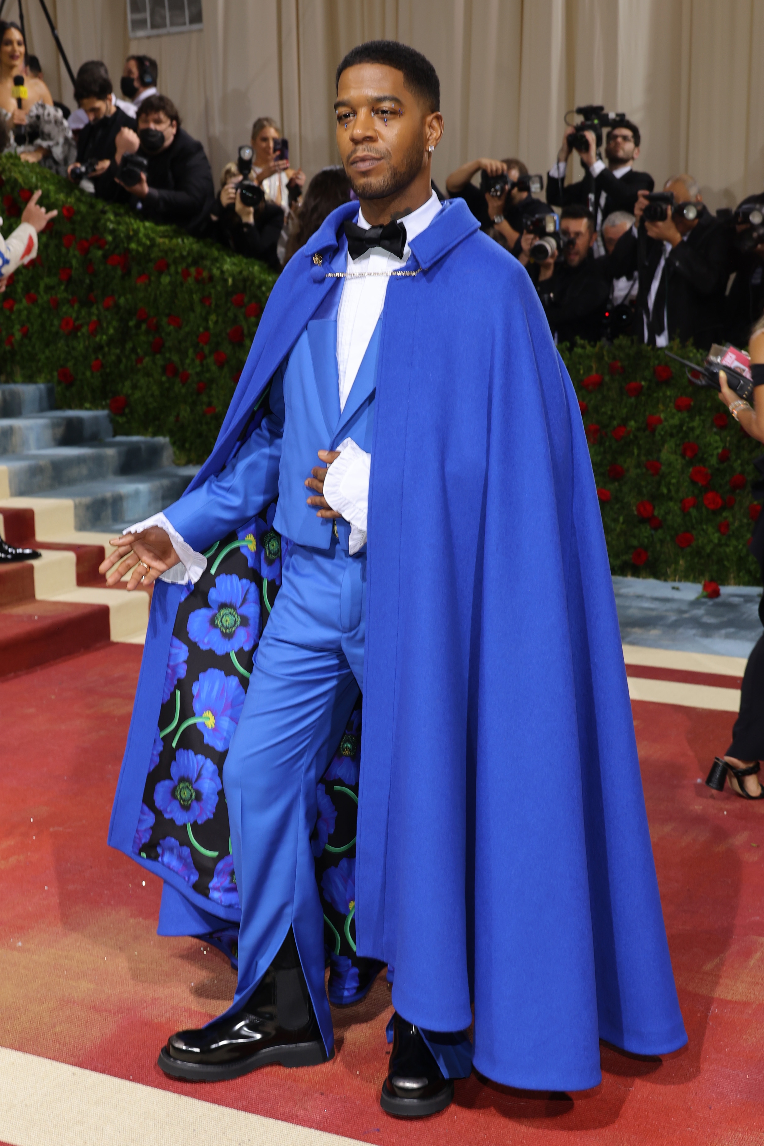 Person in a stylish suit and dramatic cape poses on a red carpet surrounded by photographers