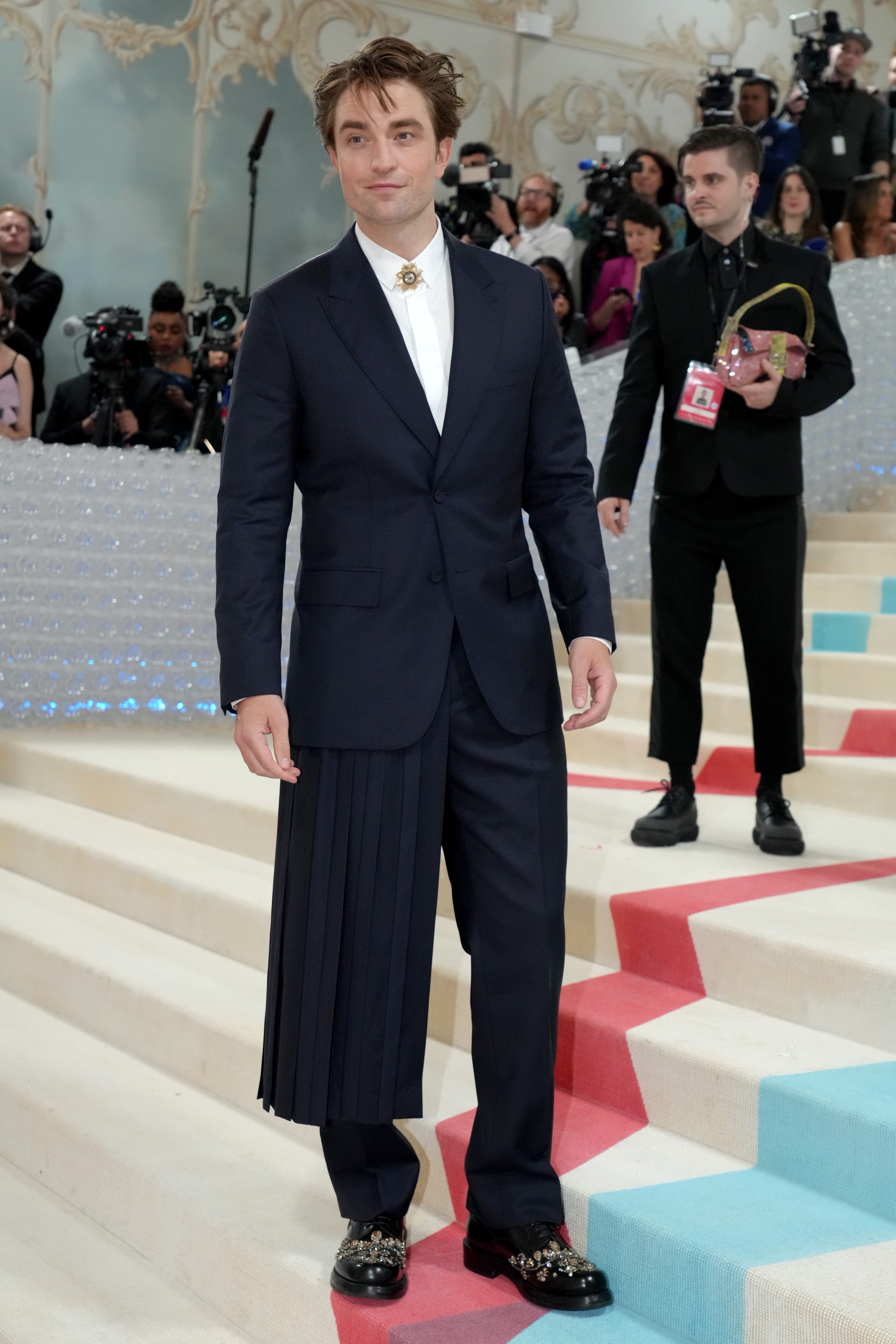 Person in a sleek, tailored suit with a unique pleated side detail, standing on a red carpet with photographers in the background