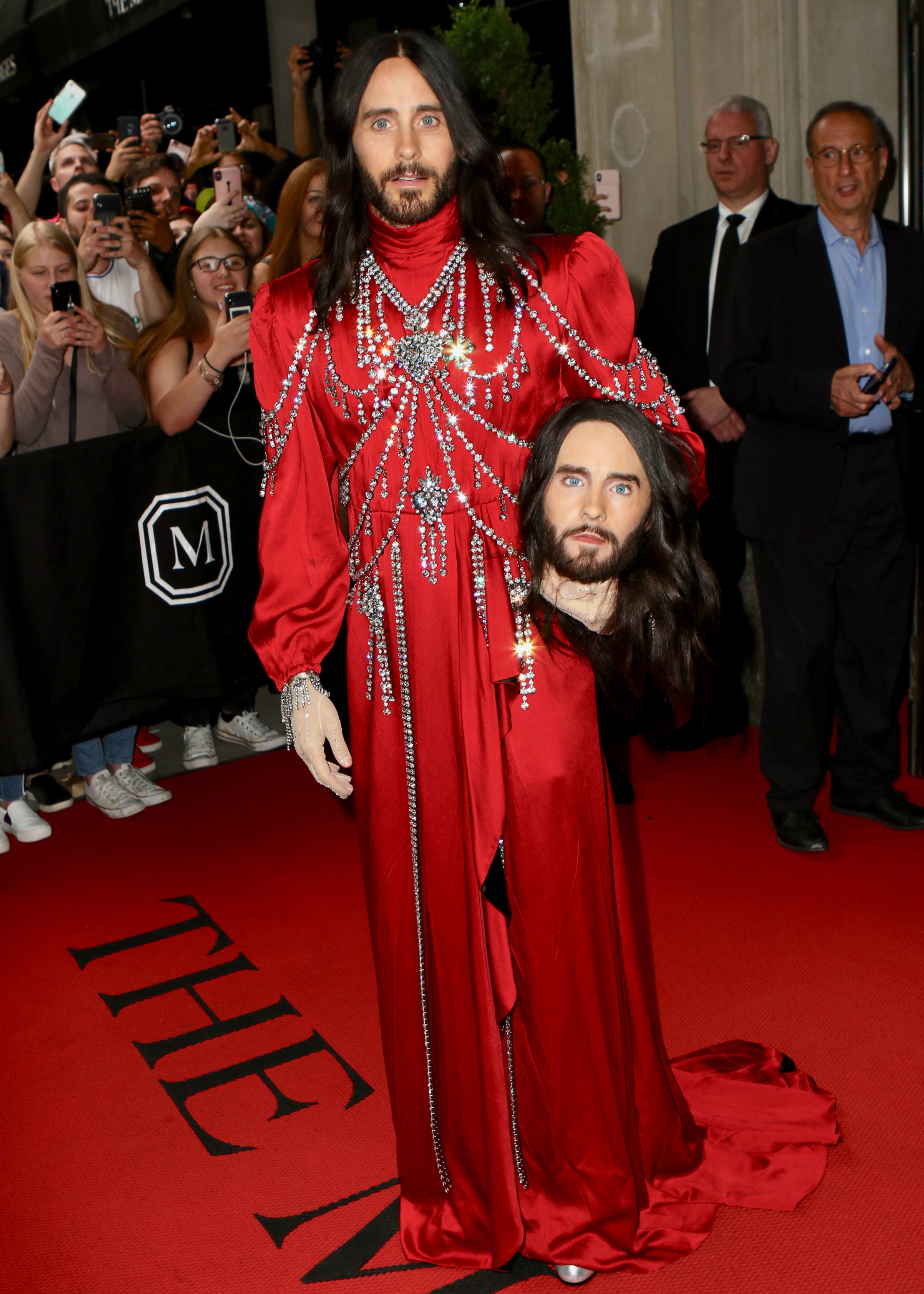 Person on red carpet in ornate red gown, holding replica of their own head. Audience in background taking photos