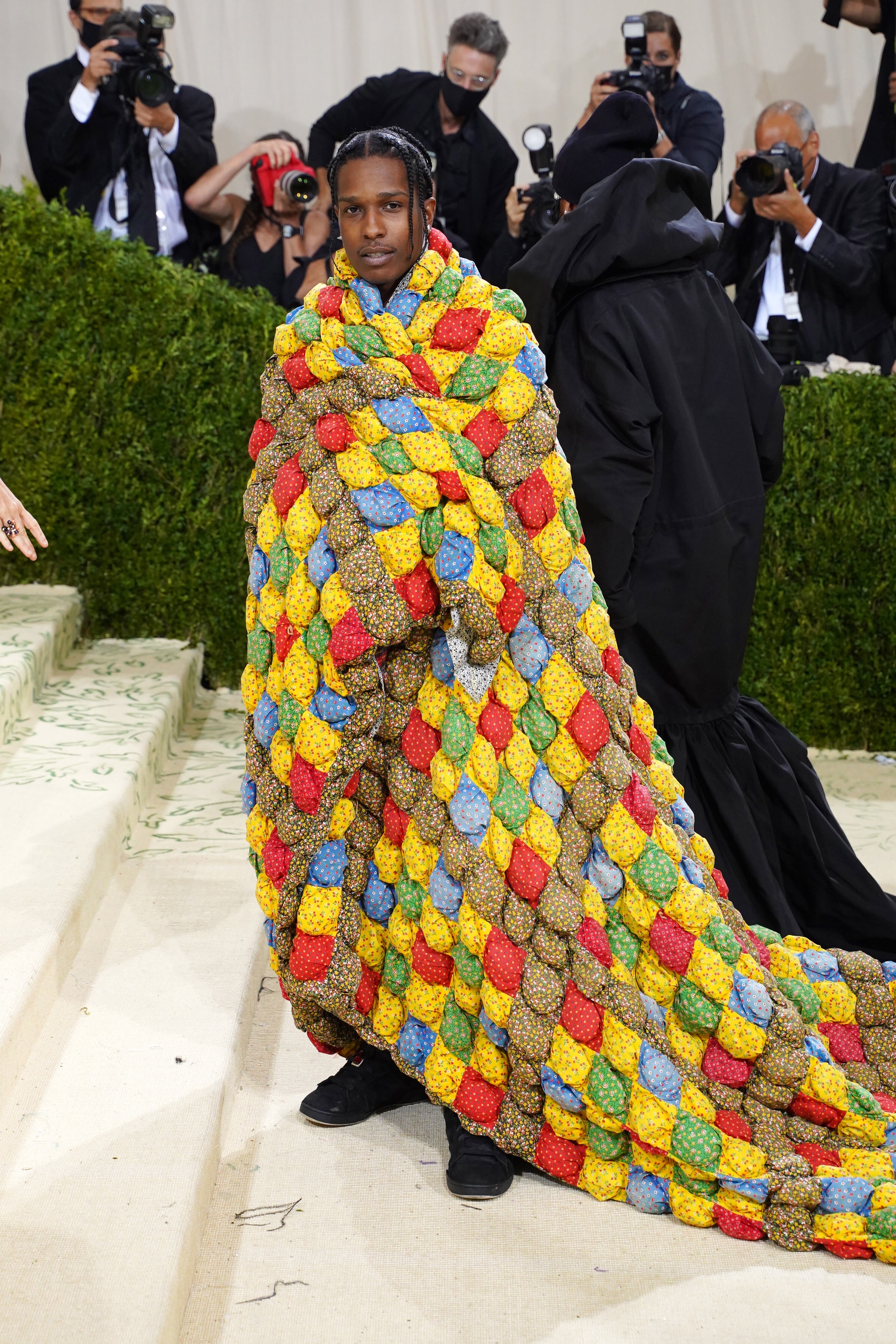 Person on a red carpet wearing a voluminous, textured quilt-like cape. Photographers in background capturing the moment