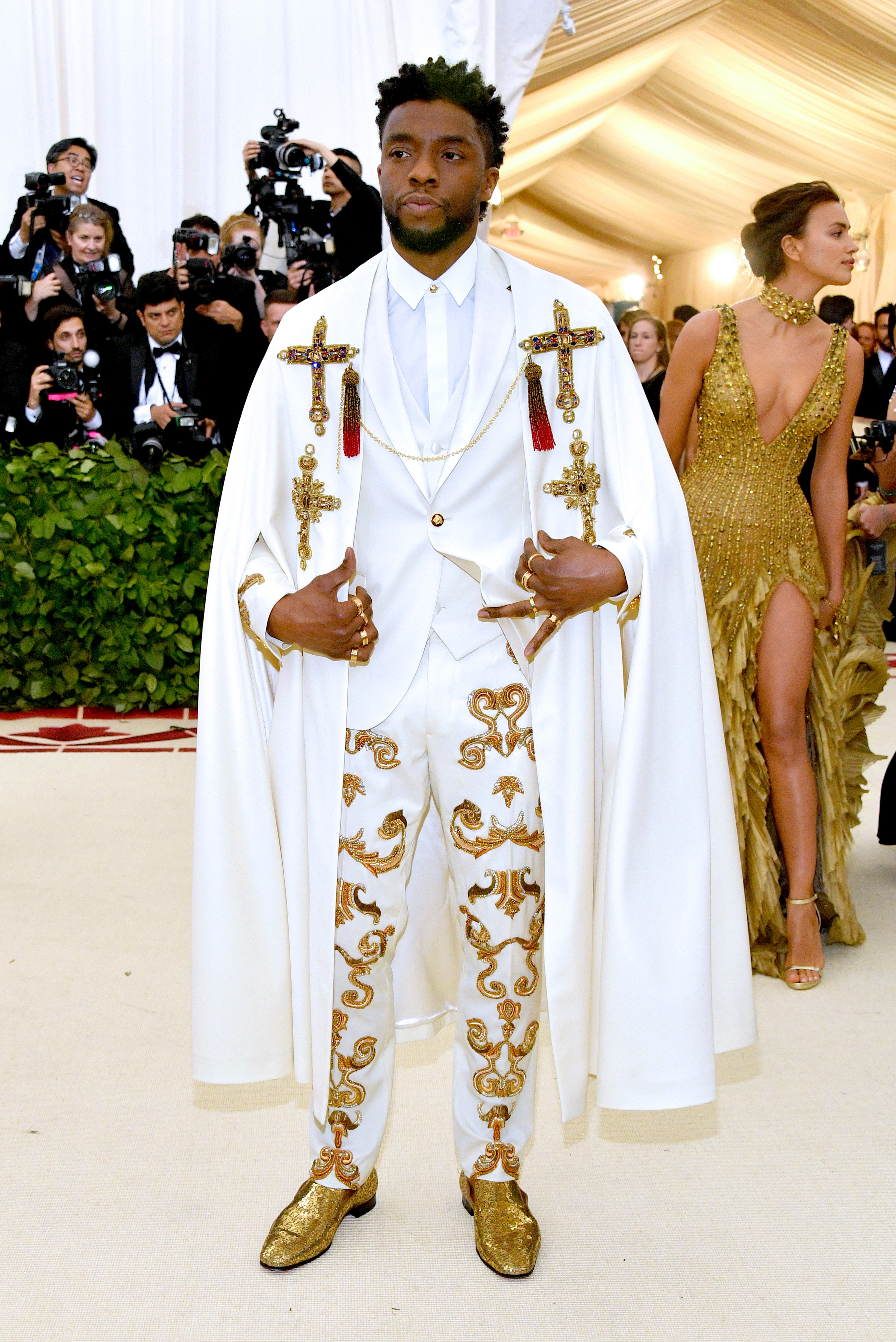 Actor in an ornate white suit with gold embroidery and a cape, posing on a red carpet. Photographers and a woman in gold dress in the background