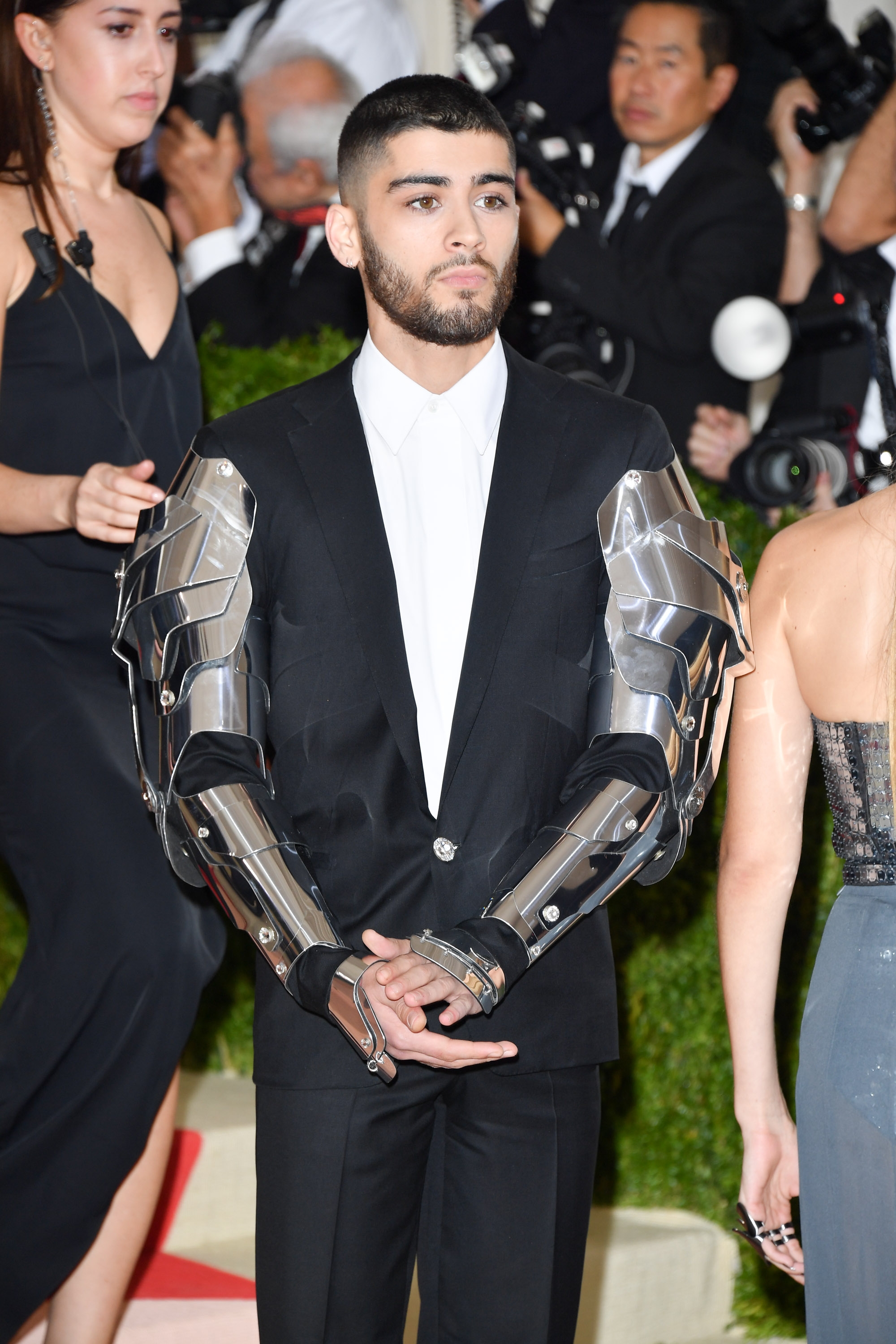A man in a suit with metallic arm accessories stands on a red carpet, surrounded by photographers