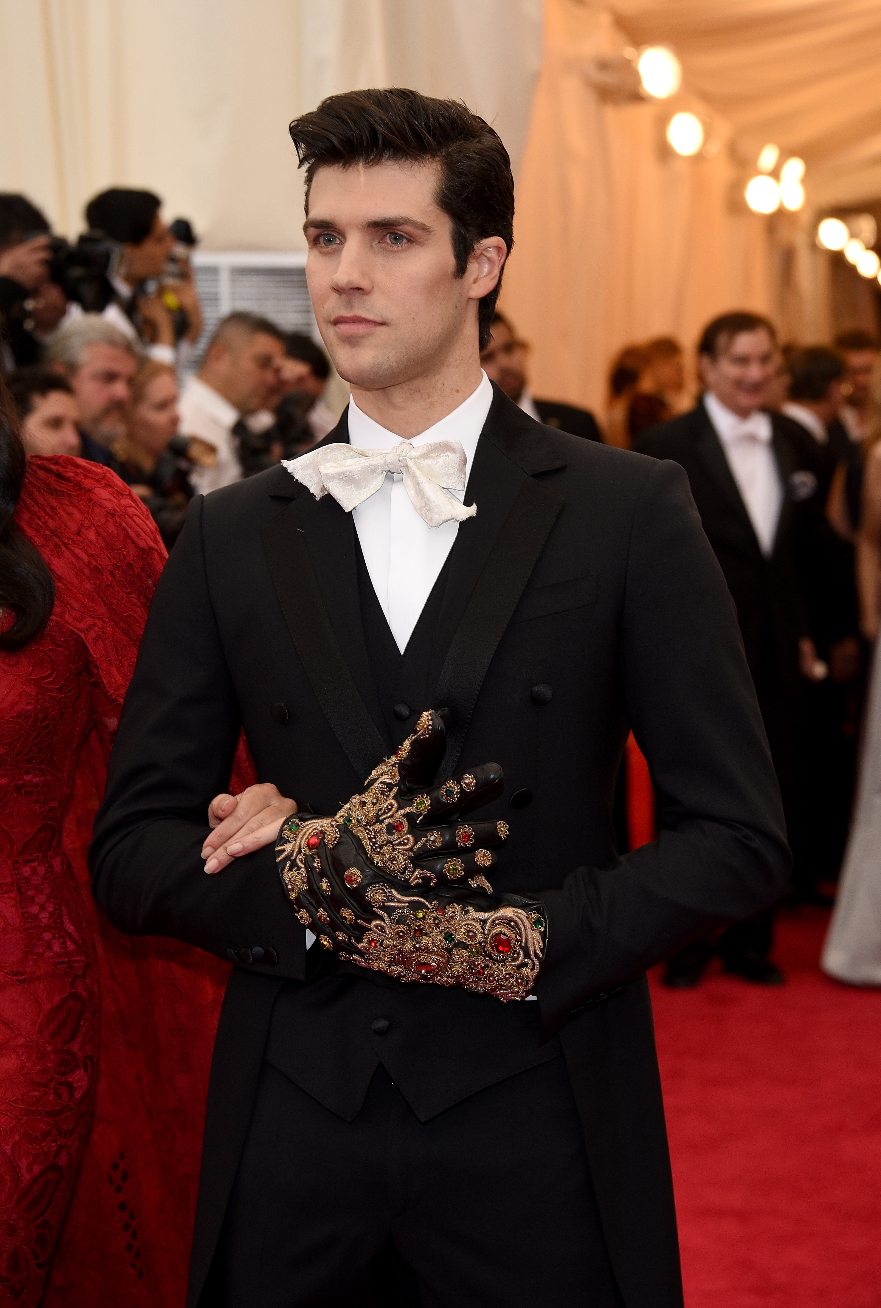 Person on red carpet in black tuxedo with a white bow tie, wearing a decorative glove with intricate details, posing for photographers