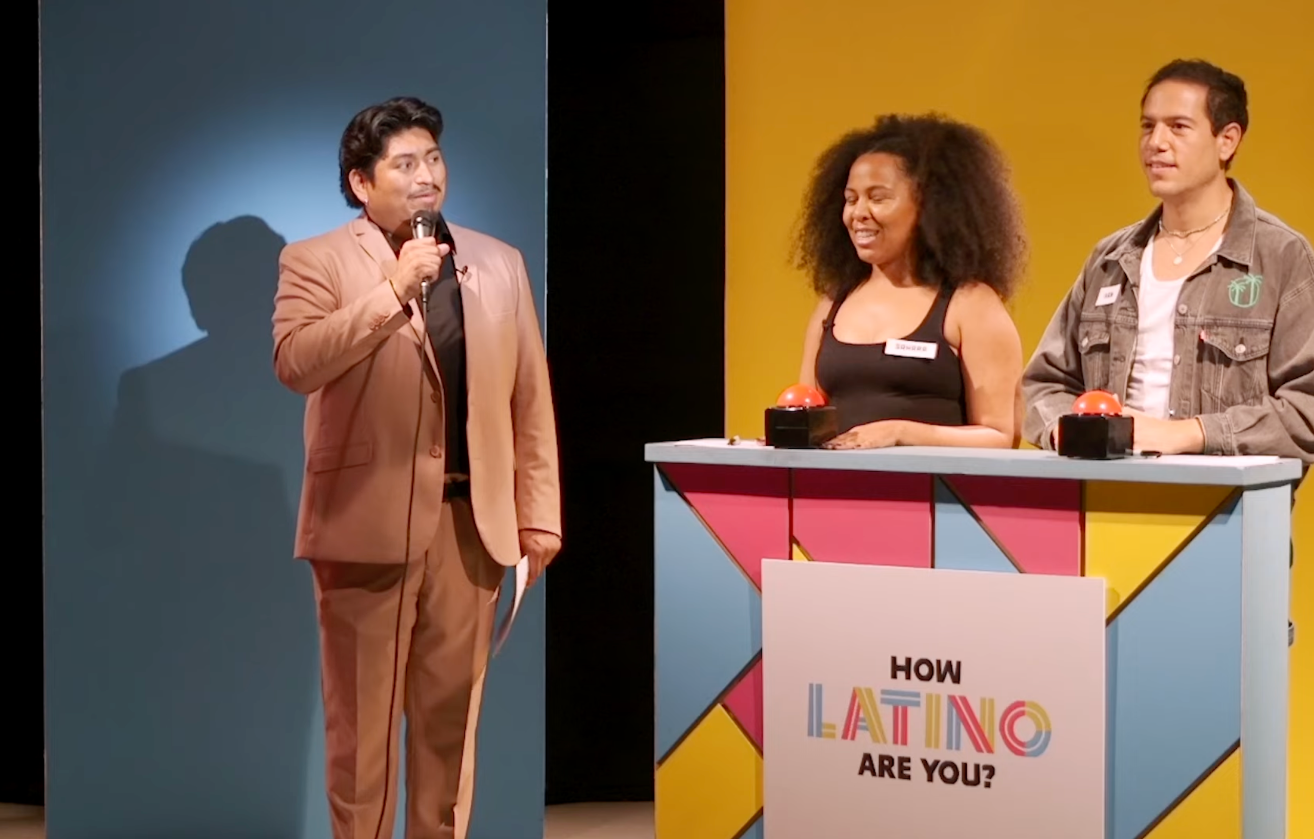 A game-show host stands with contestants at a colorful podium labeled "How Latino Are You?" with buzzers, engaging in a food-themed contest