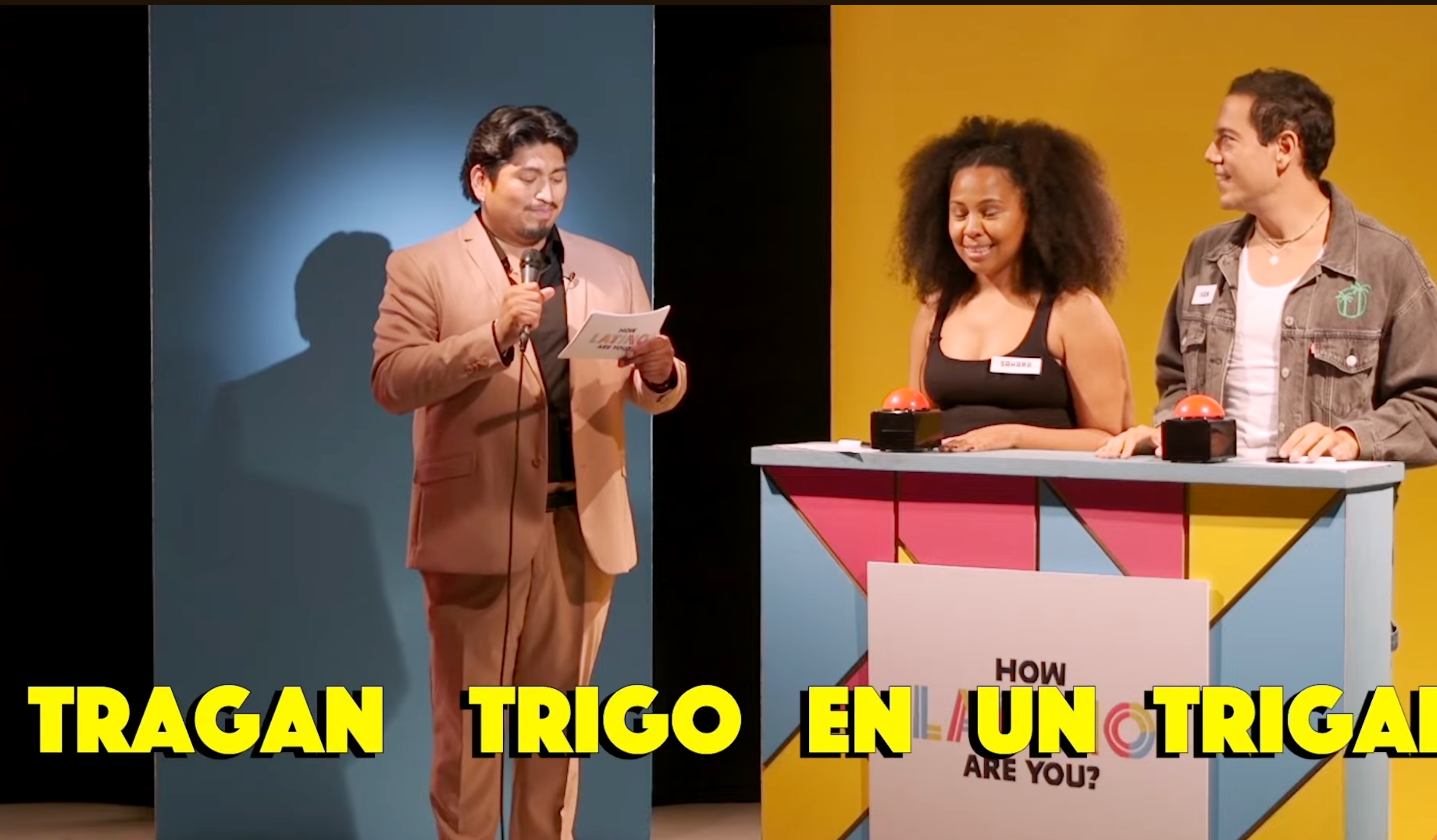 A game show host reads a card while two contestants stand at a podium, smiling. Text on screen reads "TRAGAN TRIGO EN UN TRIGAL."