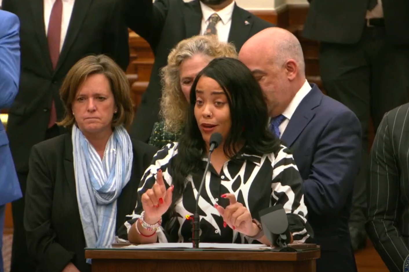 A woman stands at a podium speaking passionately, surrounded by attentive individuals in formal attire, at a public event or gathering