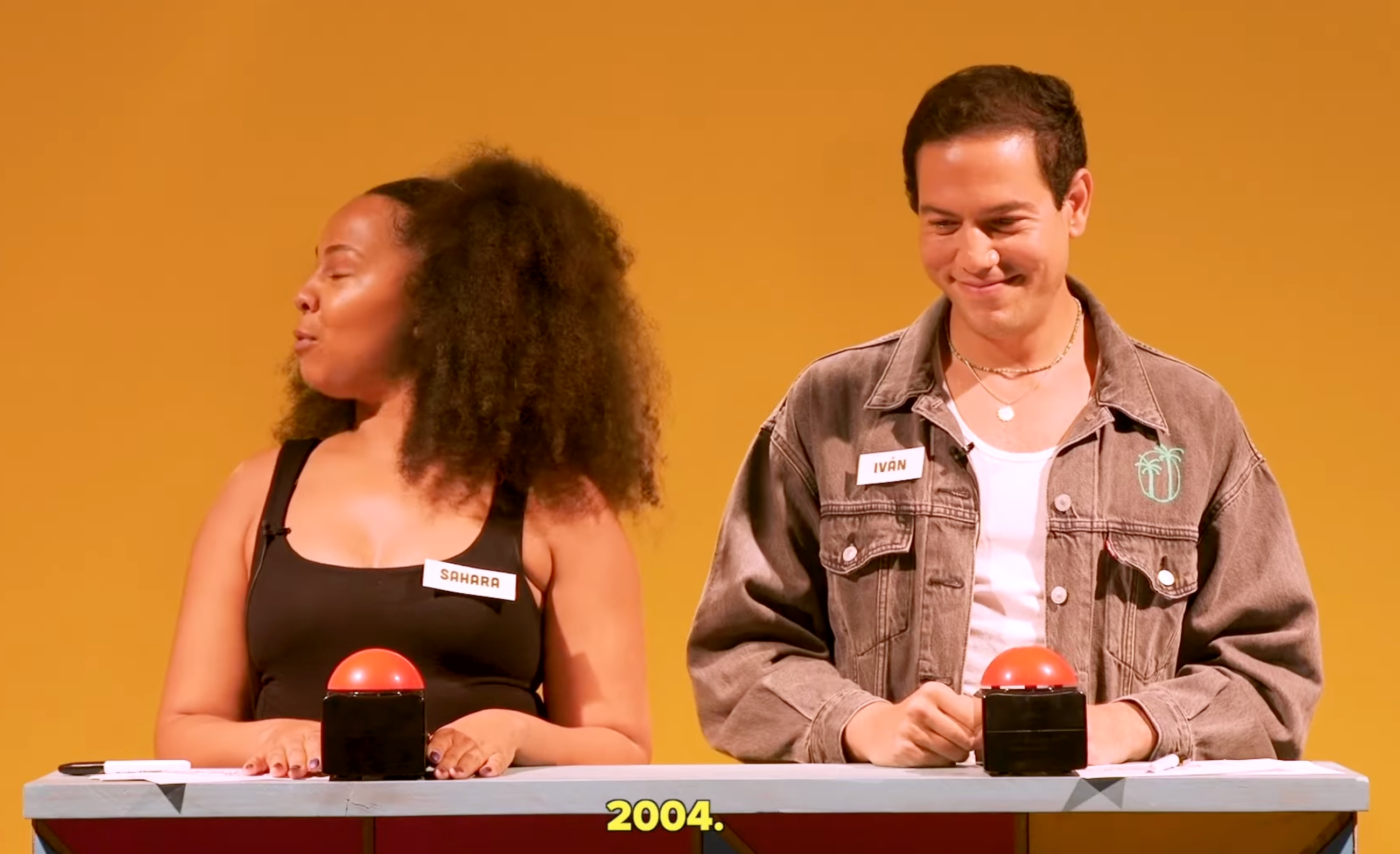 Two people stand at a game show podium with name tags. Each has a red button. The year "2004" is displayed below them