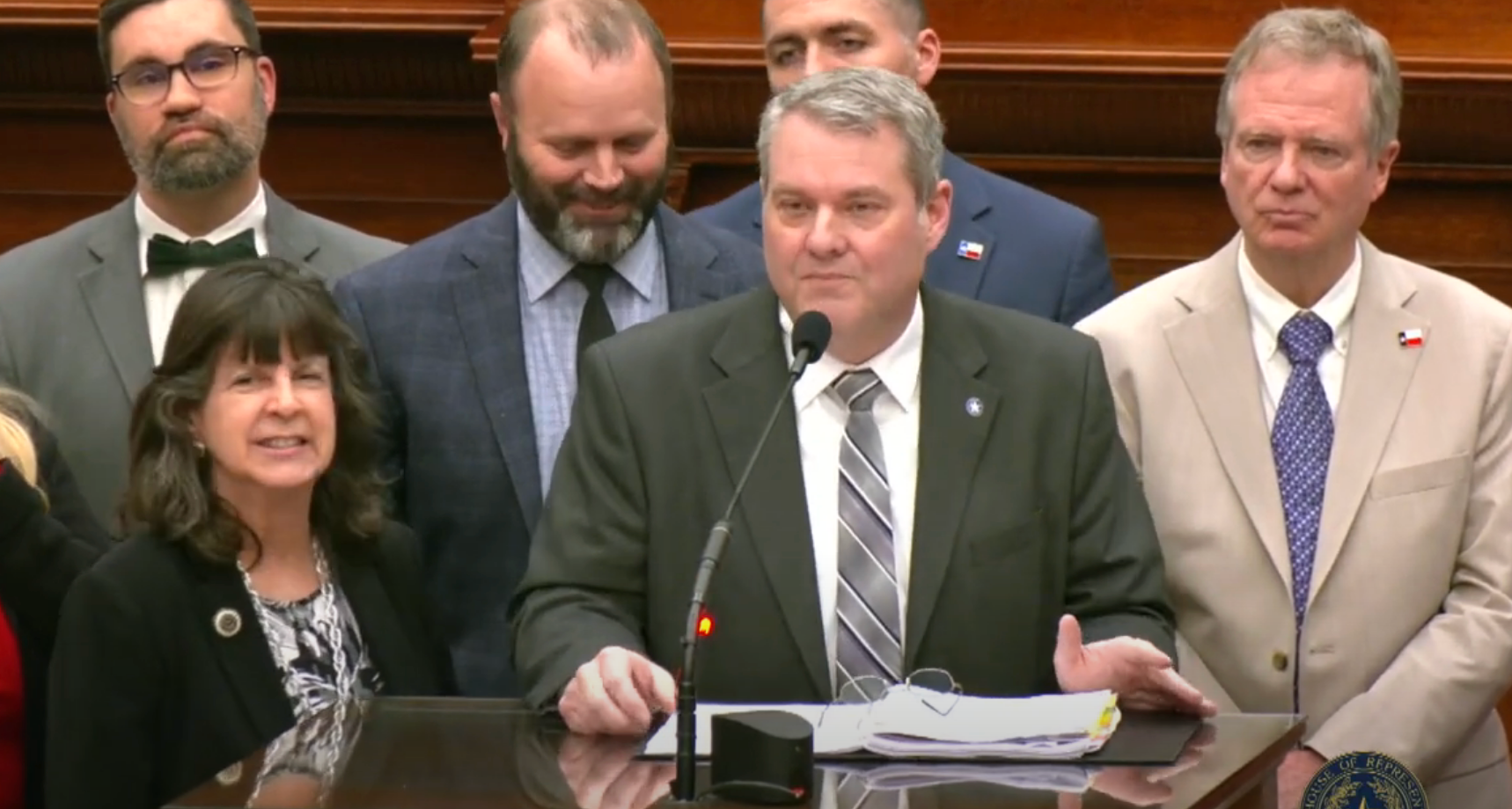 Group of people standing at a podium, one person speaking, others smiling, likely at a formal event or announcement