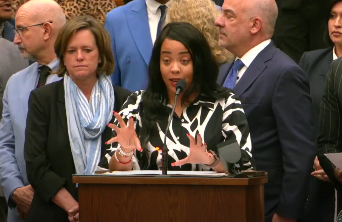 Person speaking passionately at a podium surrounded by formally dressed individuals at a press event