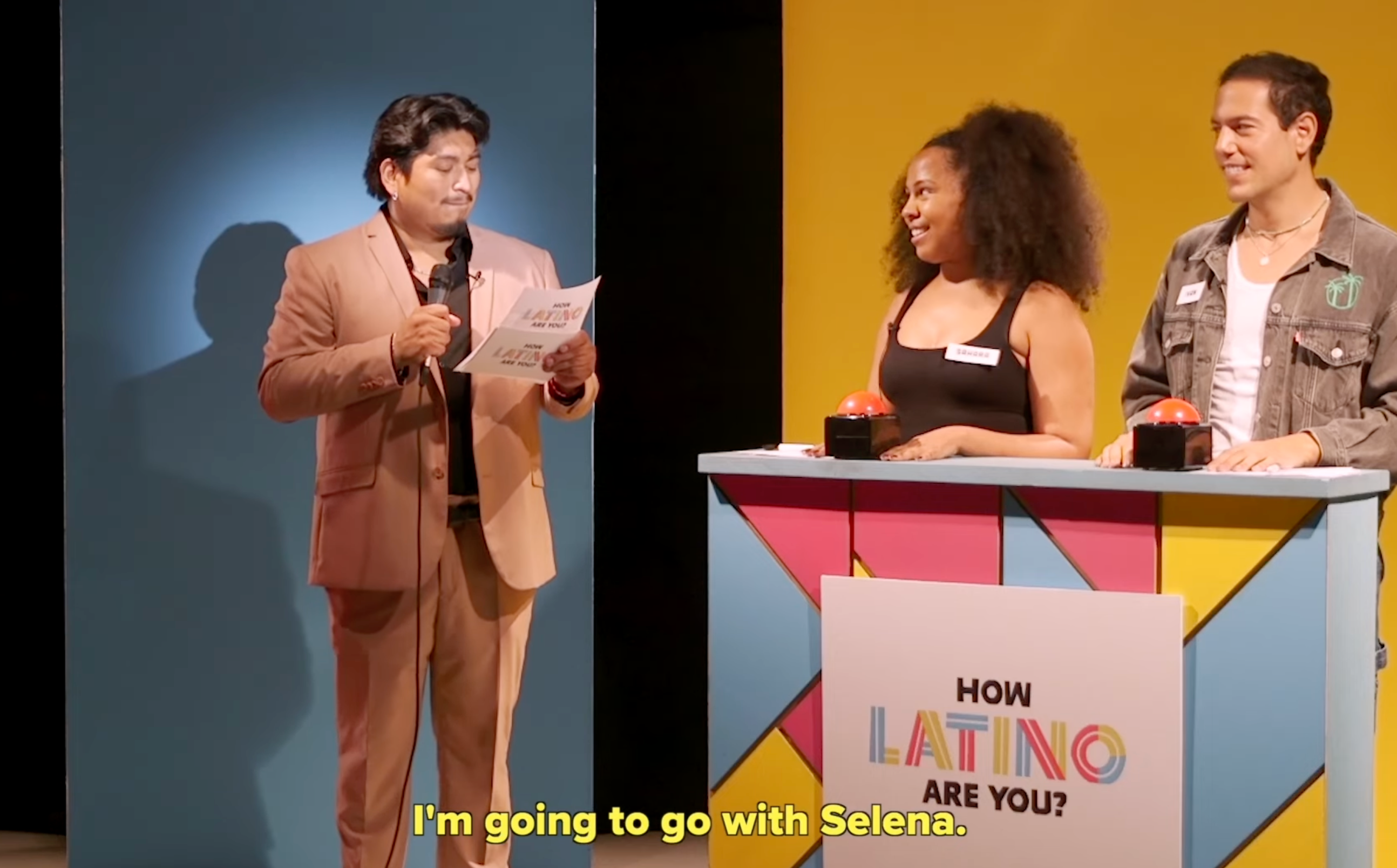 A person in a suit hosts a quiz show. Two contestants stand behind a podium with text that reads, "How Latino Are You?"