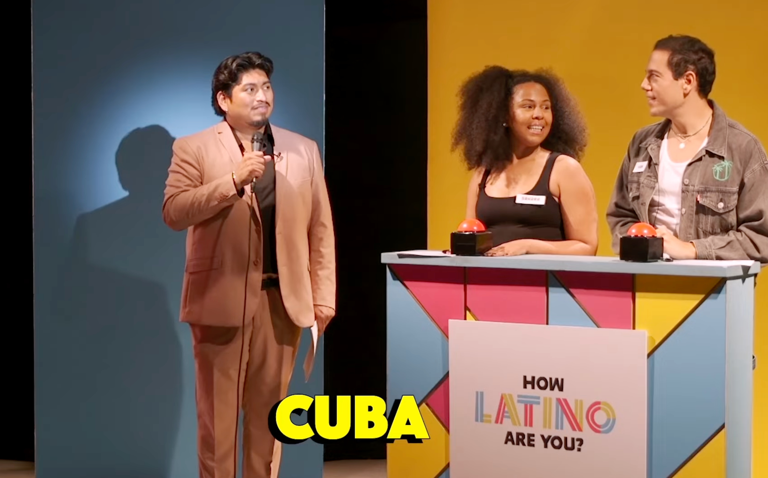 Game show scene with host and two contestants, one male and one female, standing by a podium. Onscreen text reads "CUBA"