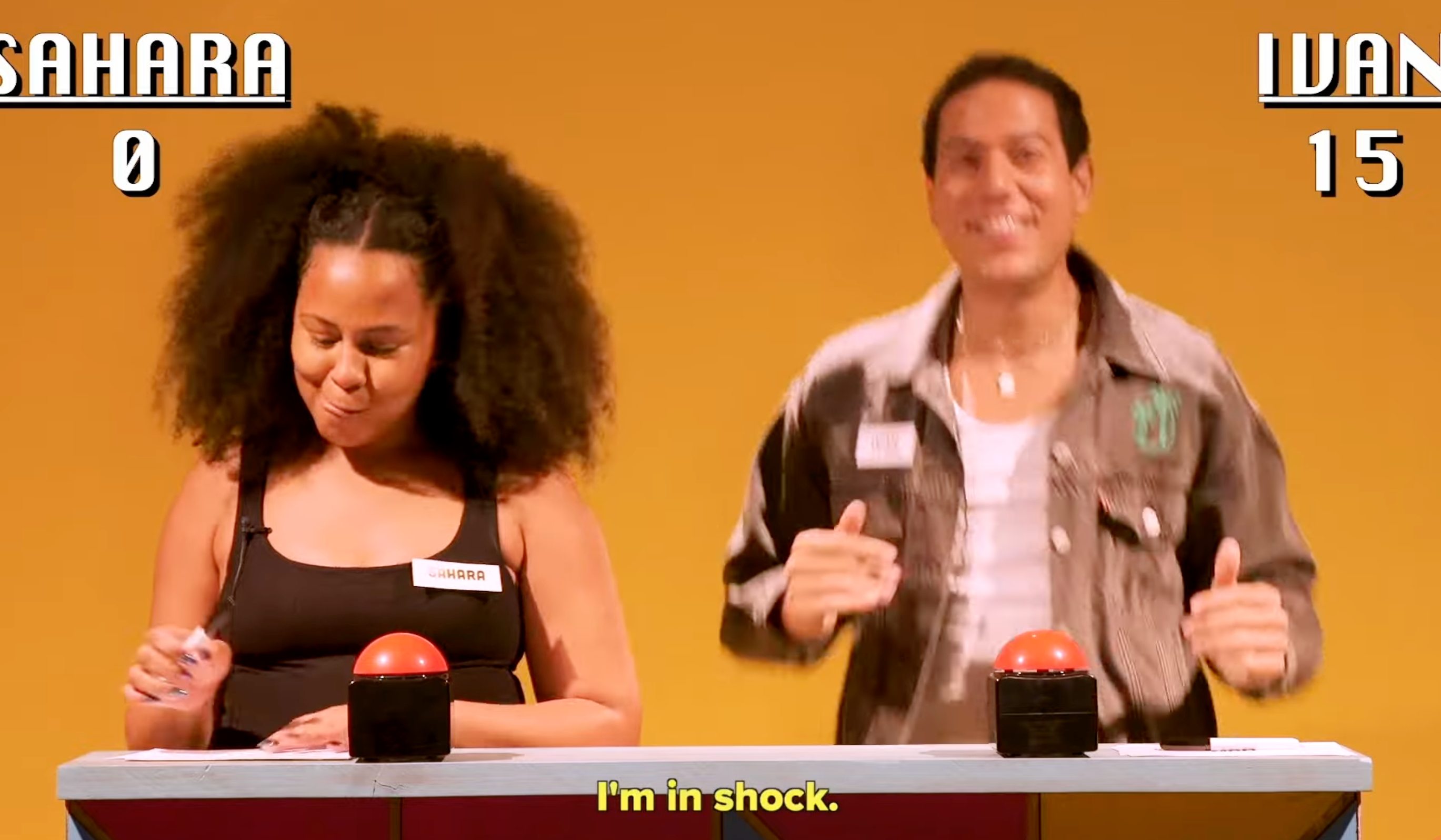 Two people in a game show setting with buzzers; one appears pleased, the other focused. Subtitles read: "I'm in shock."