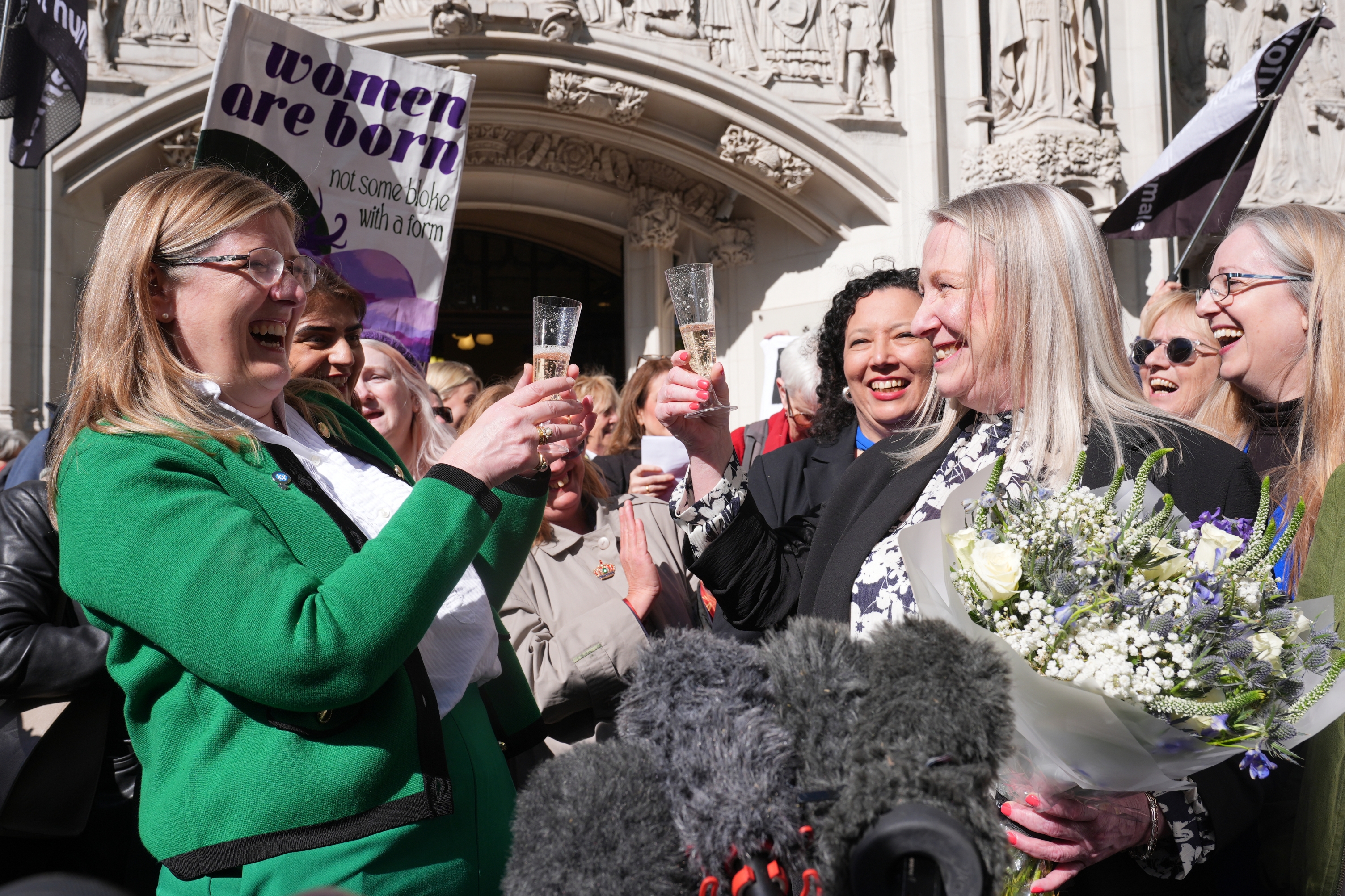 Group of people celebrating outside a building, holding champagne and flowers, with visible text on a sign reading &quot;women are born.&quot;