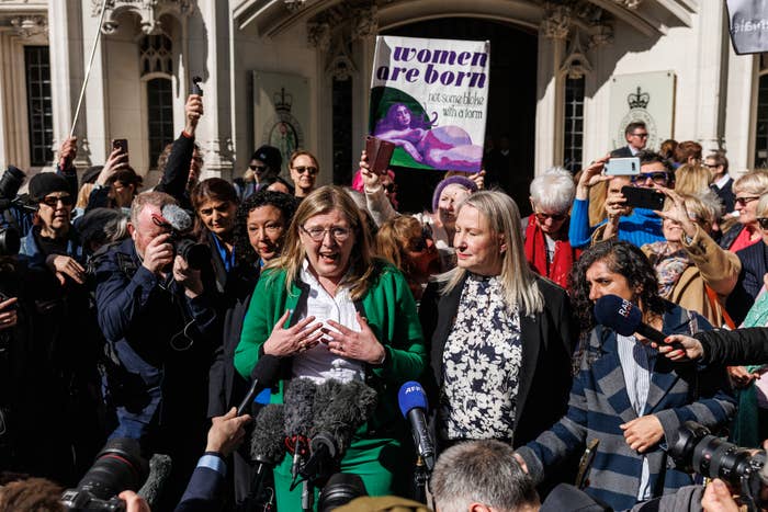 Two women, one in a green suit and the other in black-and-white, speak to the press amidst a crowd. A protester holds a &quot;women are born&quot; sign