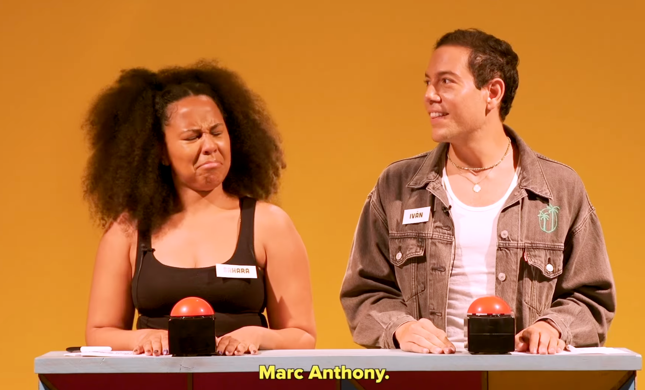 Two people stand behind a game show podium with buzzer buttons. A woman reacts animatedly while a man looks at her and smiles. Text: "Marc Anthony."