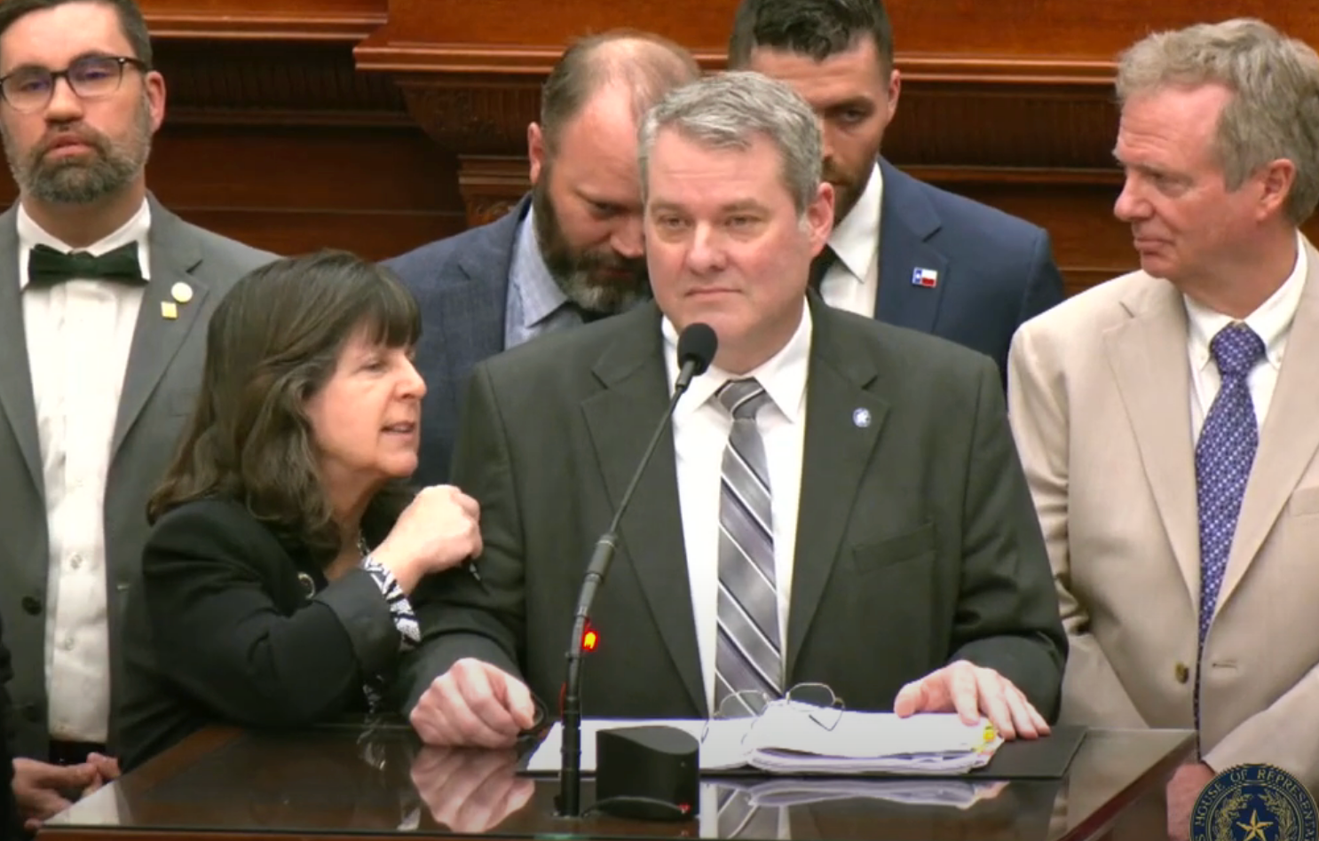 Group of formally dressed individuals stand behind a podium during a press conference, with one woman speaking while others listen attentively