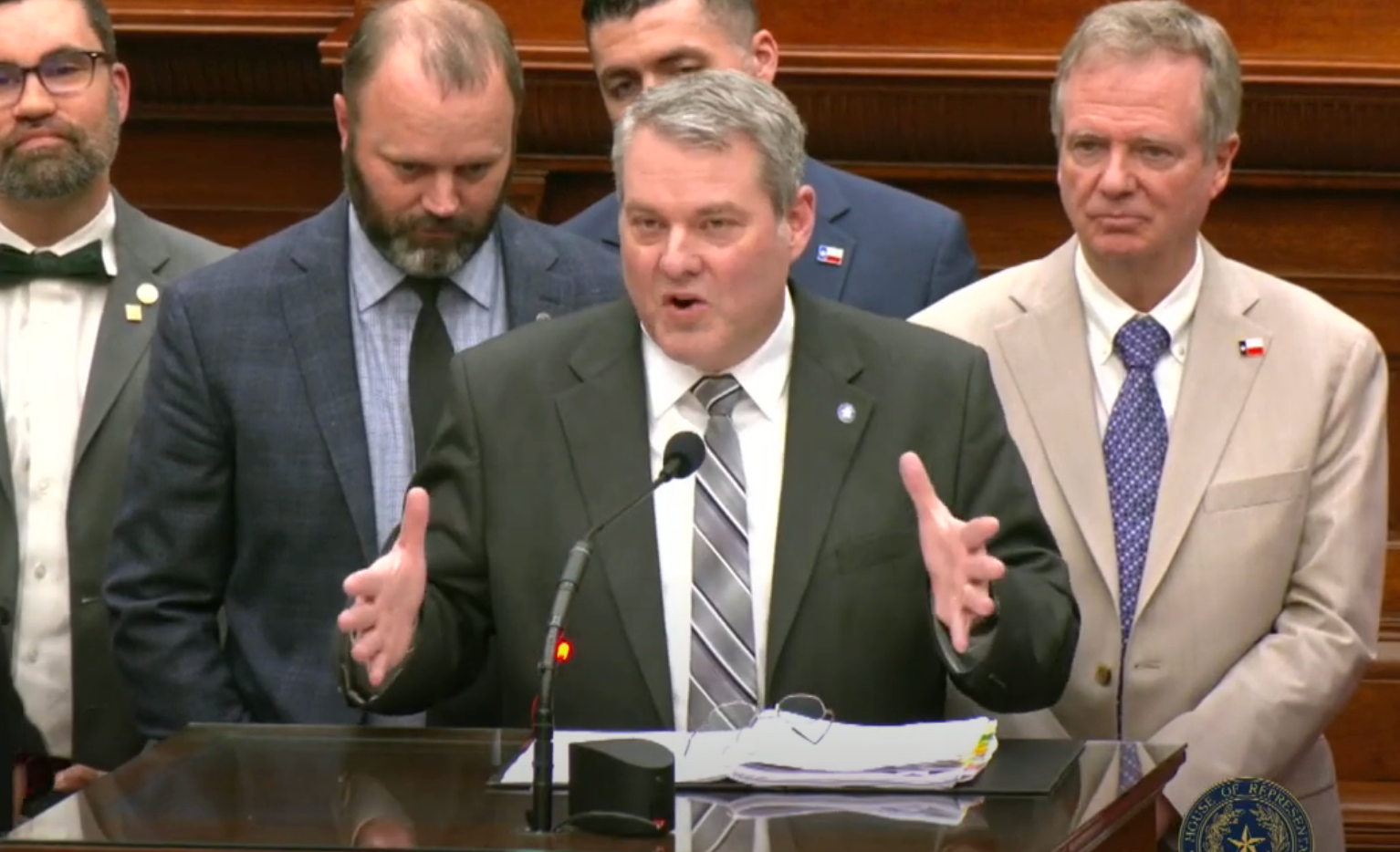 A group of suited individuals stands at a podium. A man in a suit and striped tie speaks, gesturing with his hands