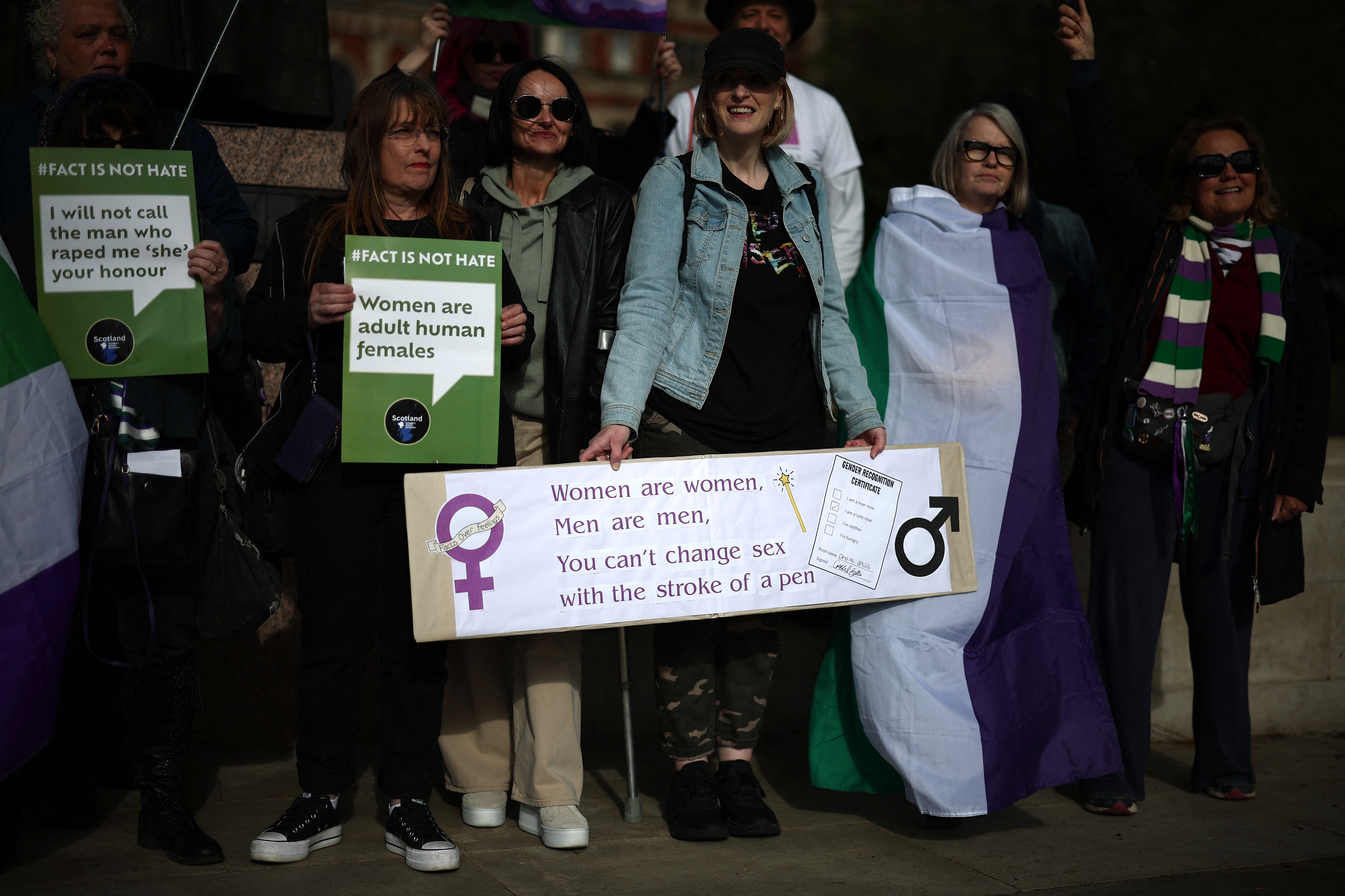 A group of people hold signs, including &quot;Women are women&quot; and LGBT flags, at a public gathering