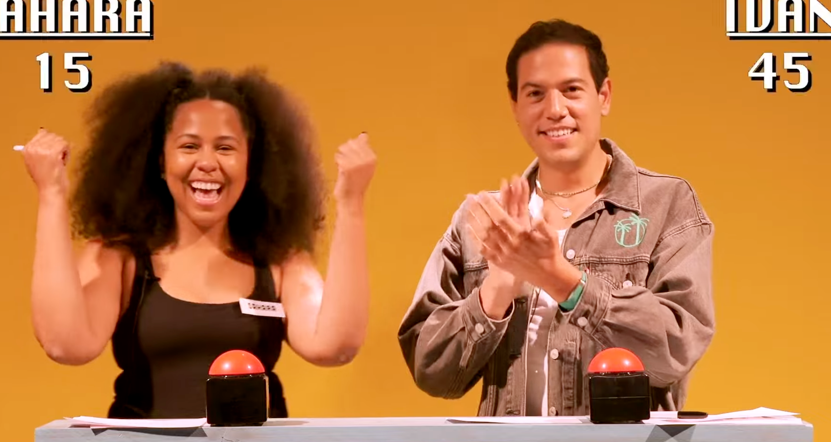 Two people stand behind game show buzzers, celebrating. The scoreboard shows Sahara with 15 points and Idan with 45 points