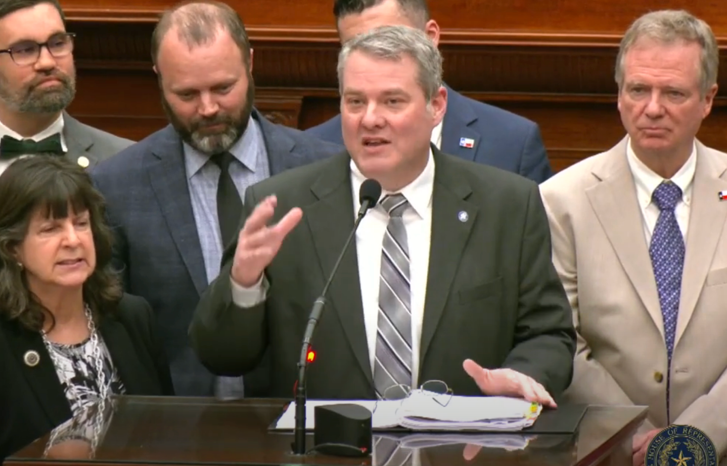 Officials stand at a podium, a speaker in a suit addresses the audience during a formal event