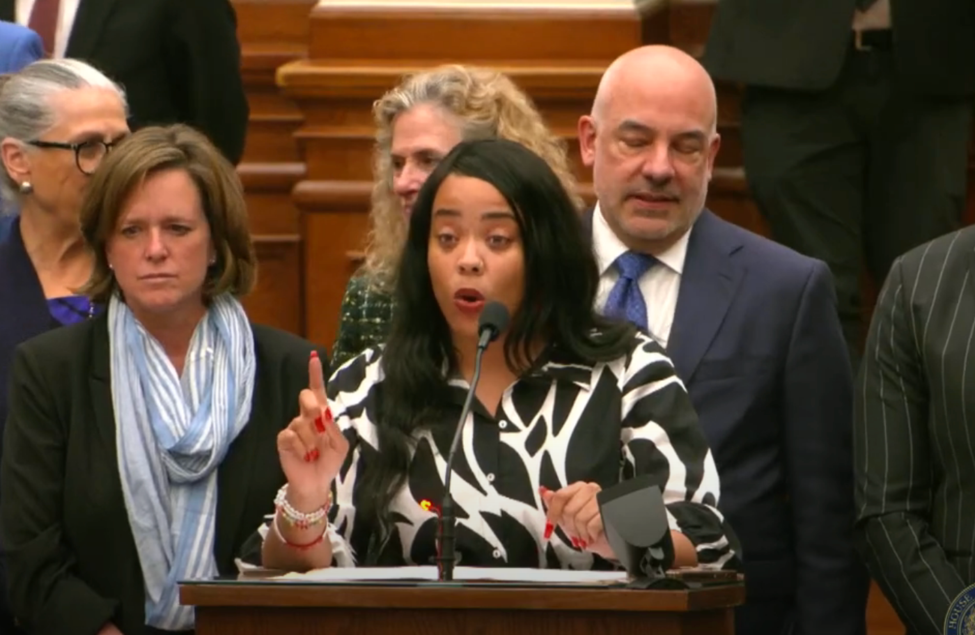 A woman speaks passionately at a podium surrounded by people in formal attire, likely during a press conference or official event