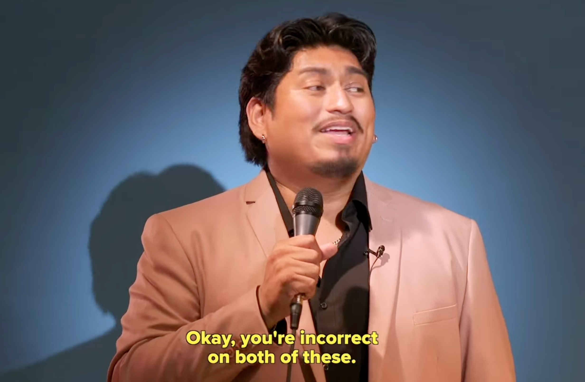 Person holding a microphone, smiling, wearing a suit jacket. Caption reads: "Okay, you're incorrect on both of these."