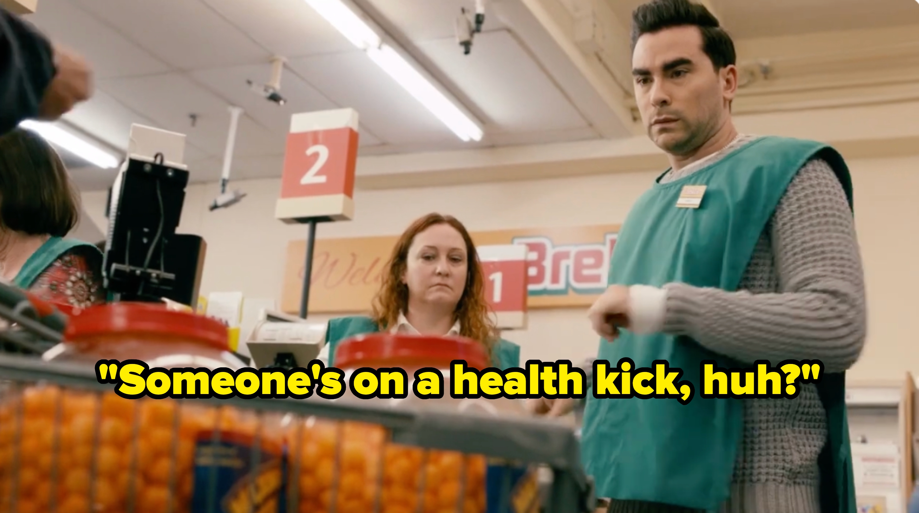 Two store employees wearing green aprons work at a checkout counter in a grocery store