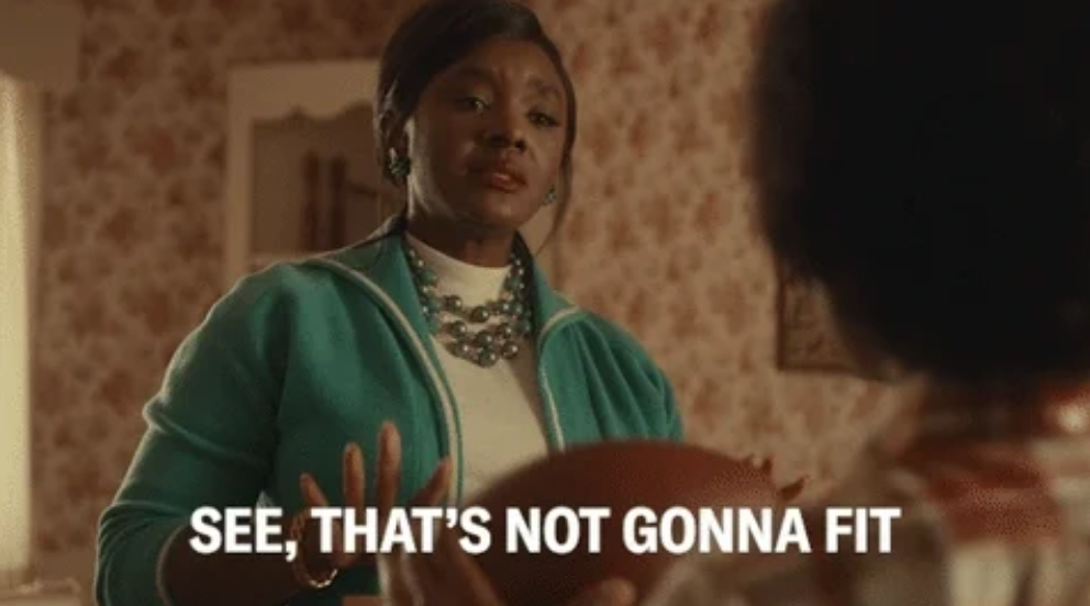 Woman holding a small football, expressing doubt to another person, with visible text: "See, that's not gonna fit."
