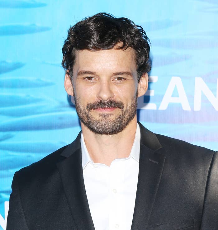 Austin Nichols in a suit with a white shirt poses in front of a blue ocean-themed backdrop