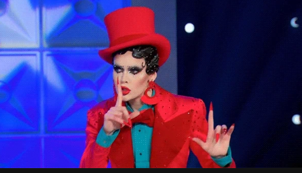 Person in theatrical costume with a red top hat, striking a dramatic pose with a finger on lips, suggesting silence