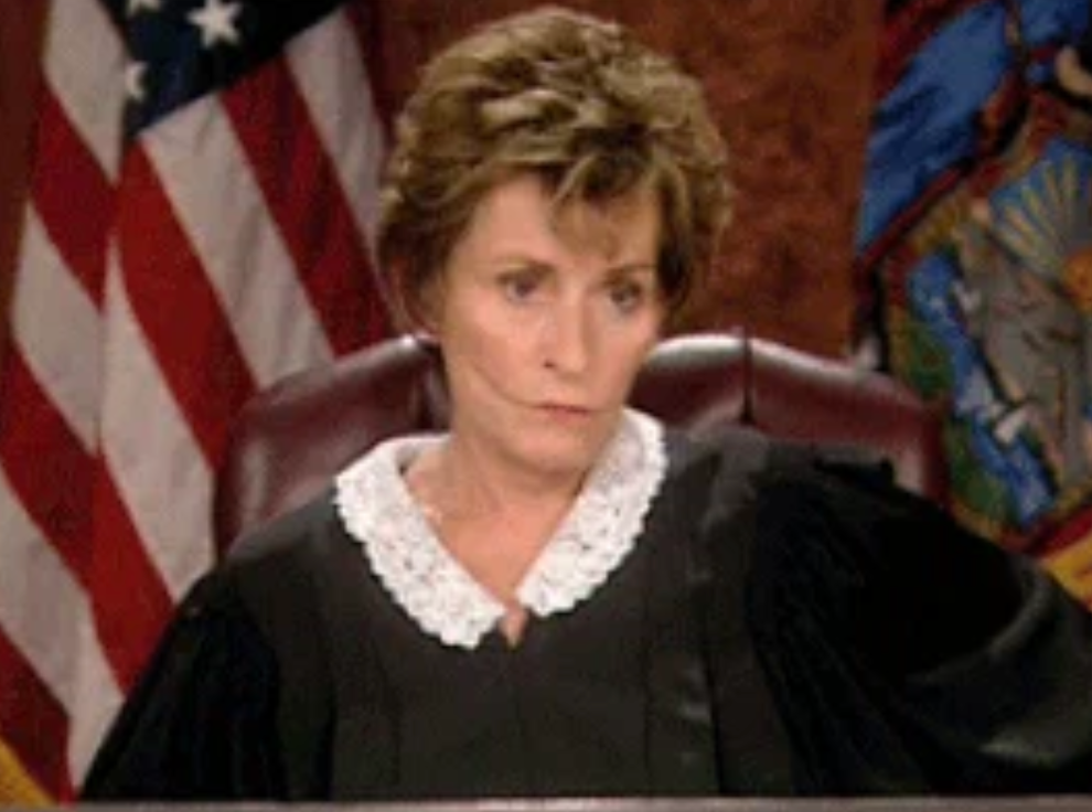 A judge in a courtroom setting, wearing a robe with a white collar, sits in front of flags, displaying a focused expression