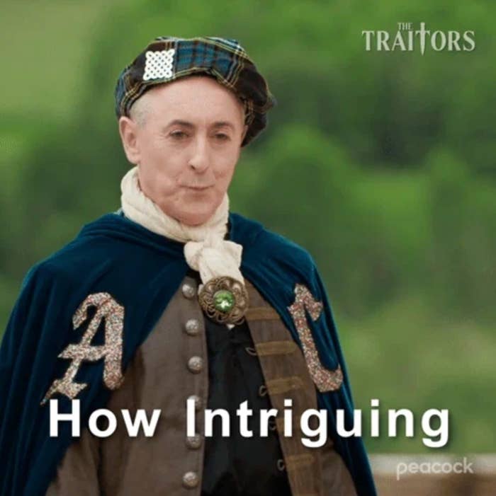 Person in historical attire with a cape and scarf, wearing a patterned hat, in a scene from "The Traitors." Text reads, "How Intriguing."