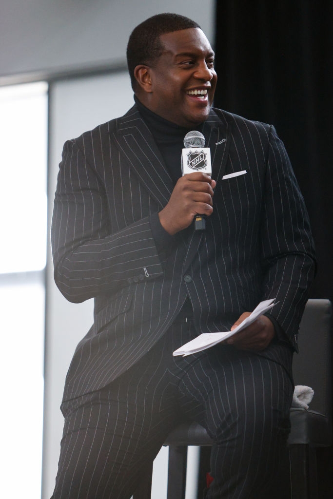 Kevin Weekes wearing a pinstripe suit, smiling while holding an NHL microphone and papers, seated on stage