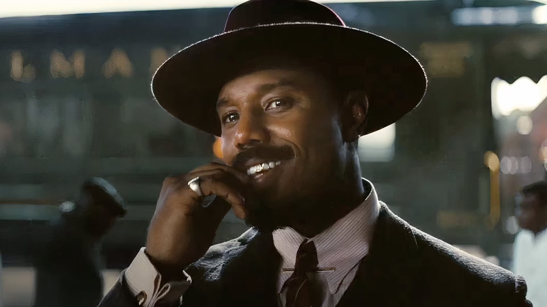 Persona sonriente con sombrero de ala ancha y traje formal, en una escena al aire libre con fondo de tren borroso