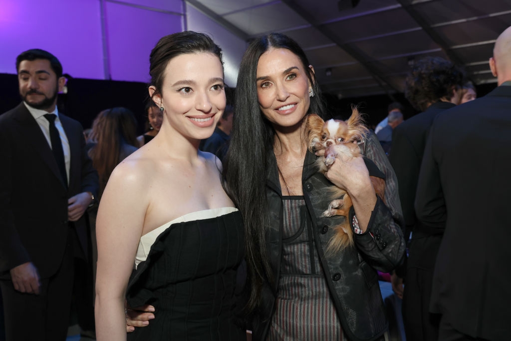 Two people pose at an event, one in a strapless dress and the other in a sheer jacket, holding a small dog
