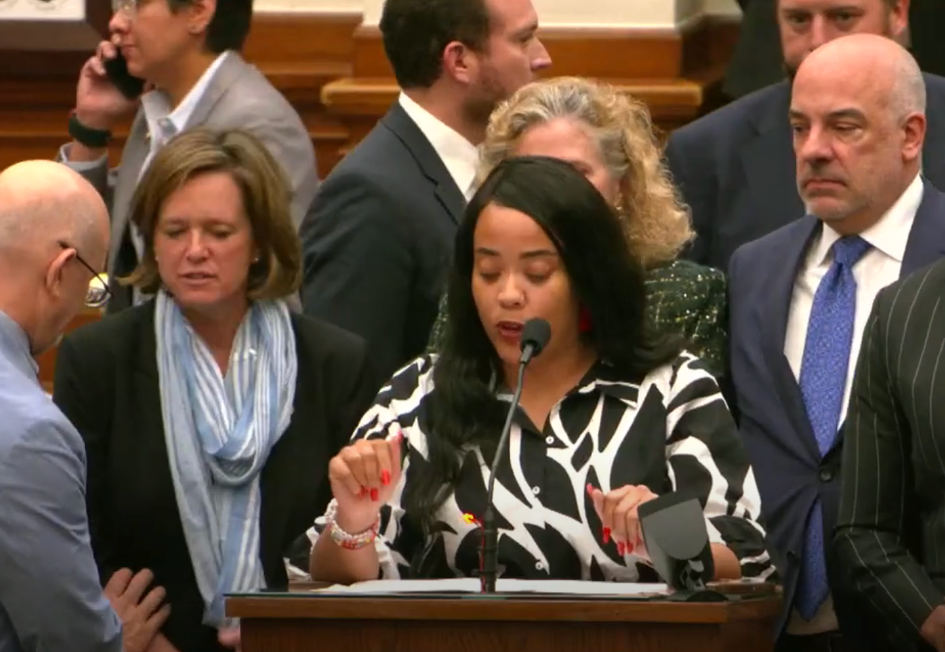 A woman speaks at a podium surrounded by people, possibly at a conference or meeting