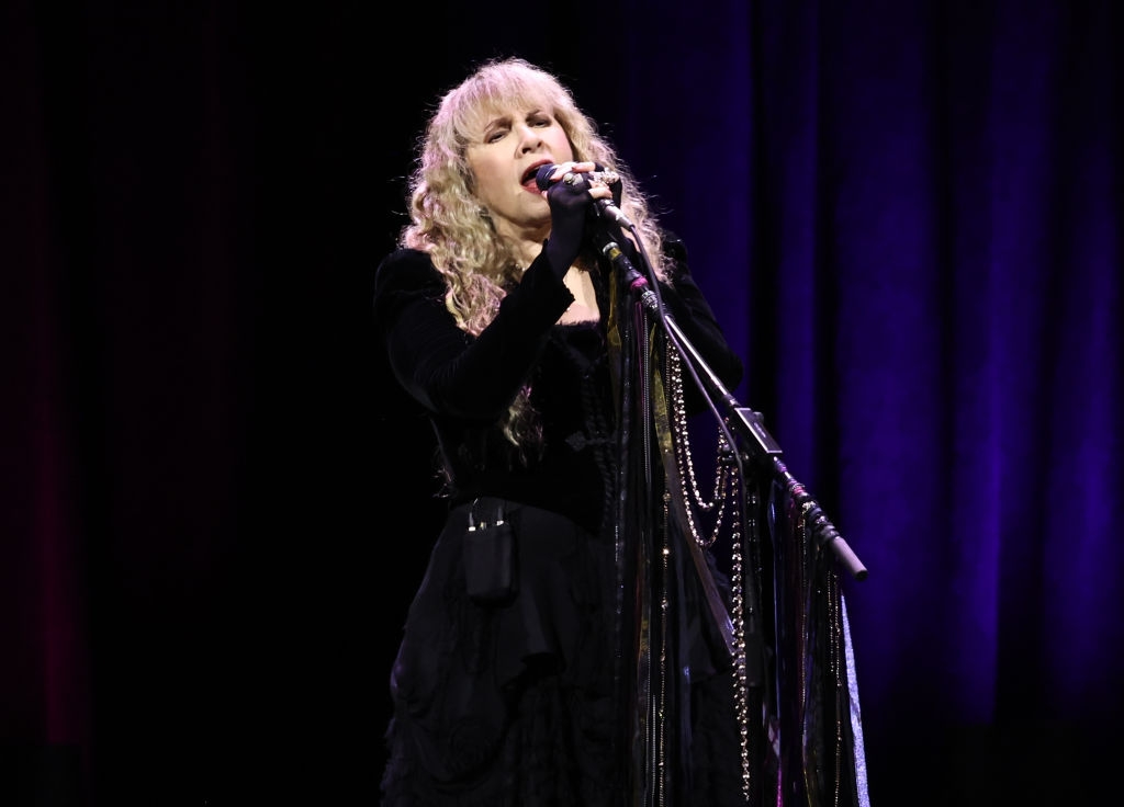 Stevie Nicks passionately performing on stage wearing a flowing black outfit with long sleeves and intricate details, holding a microphone