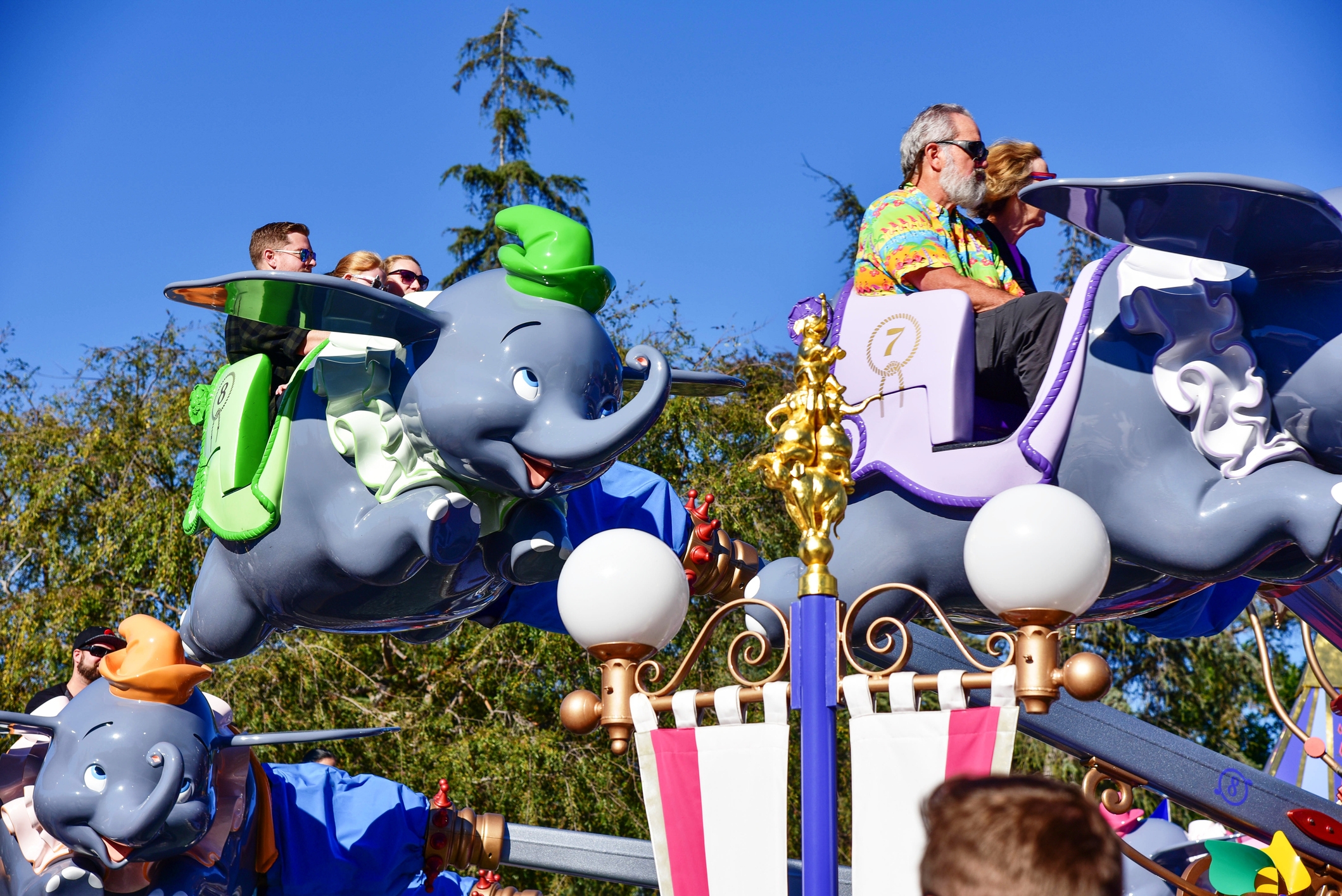 People enjoying a ride on Dumbo-themed flying elephants at an amusement park, with trees and clear sky in the background