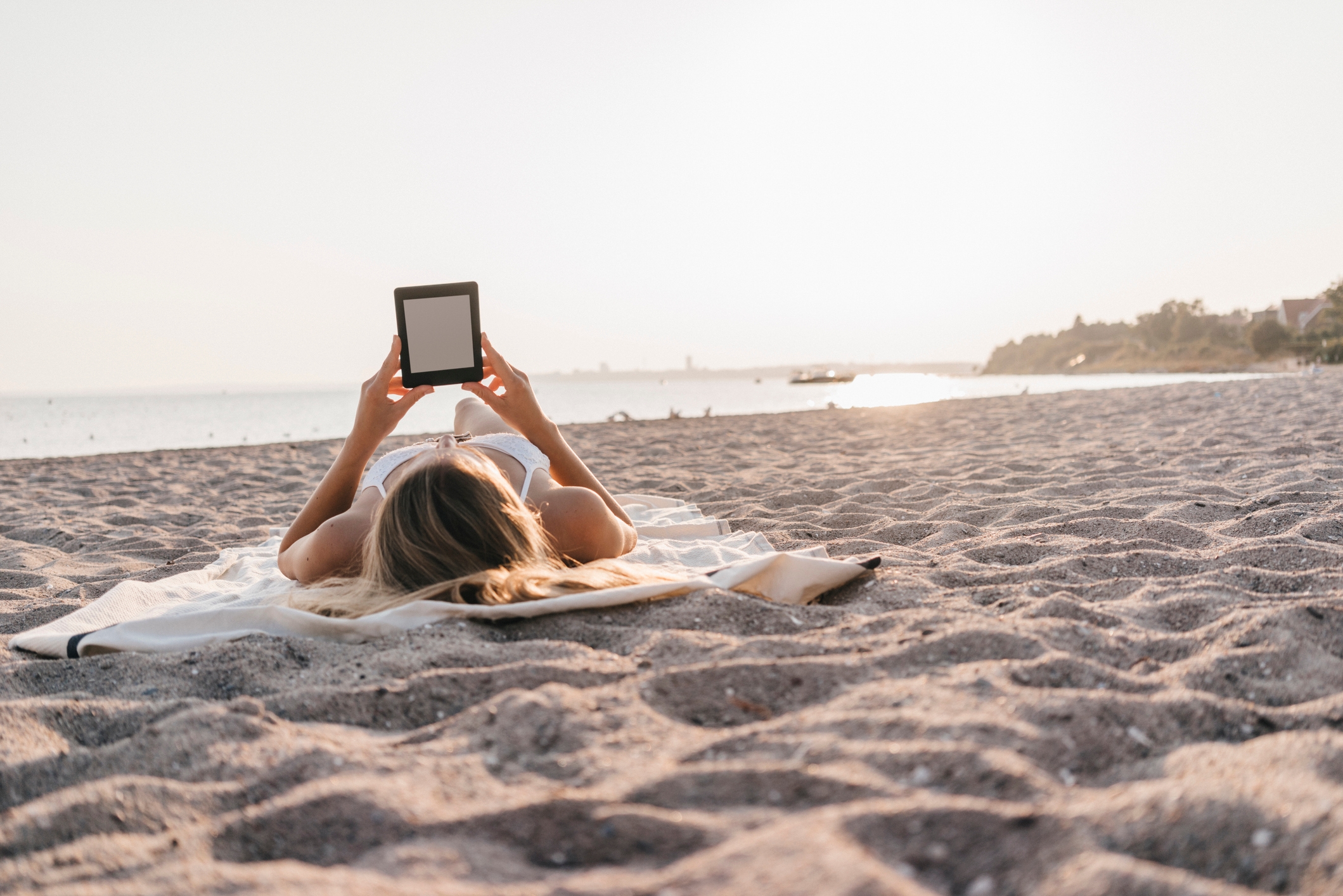 Person relaxing on a beach, lying on a towel, reading an e-reader as the sun sets over the water in the background