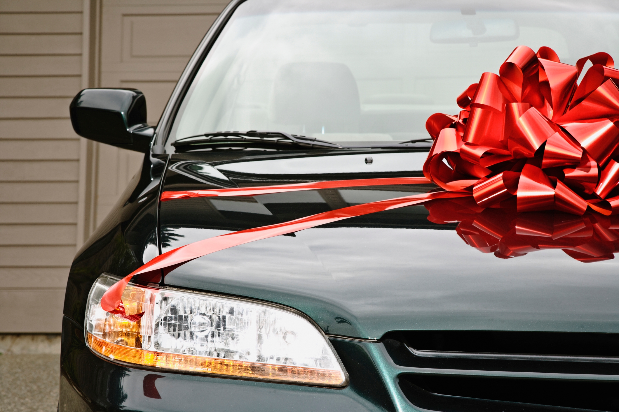 Car with a large red bow on the hood, parked in front of a garage, suggesting a gift or purchase