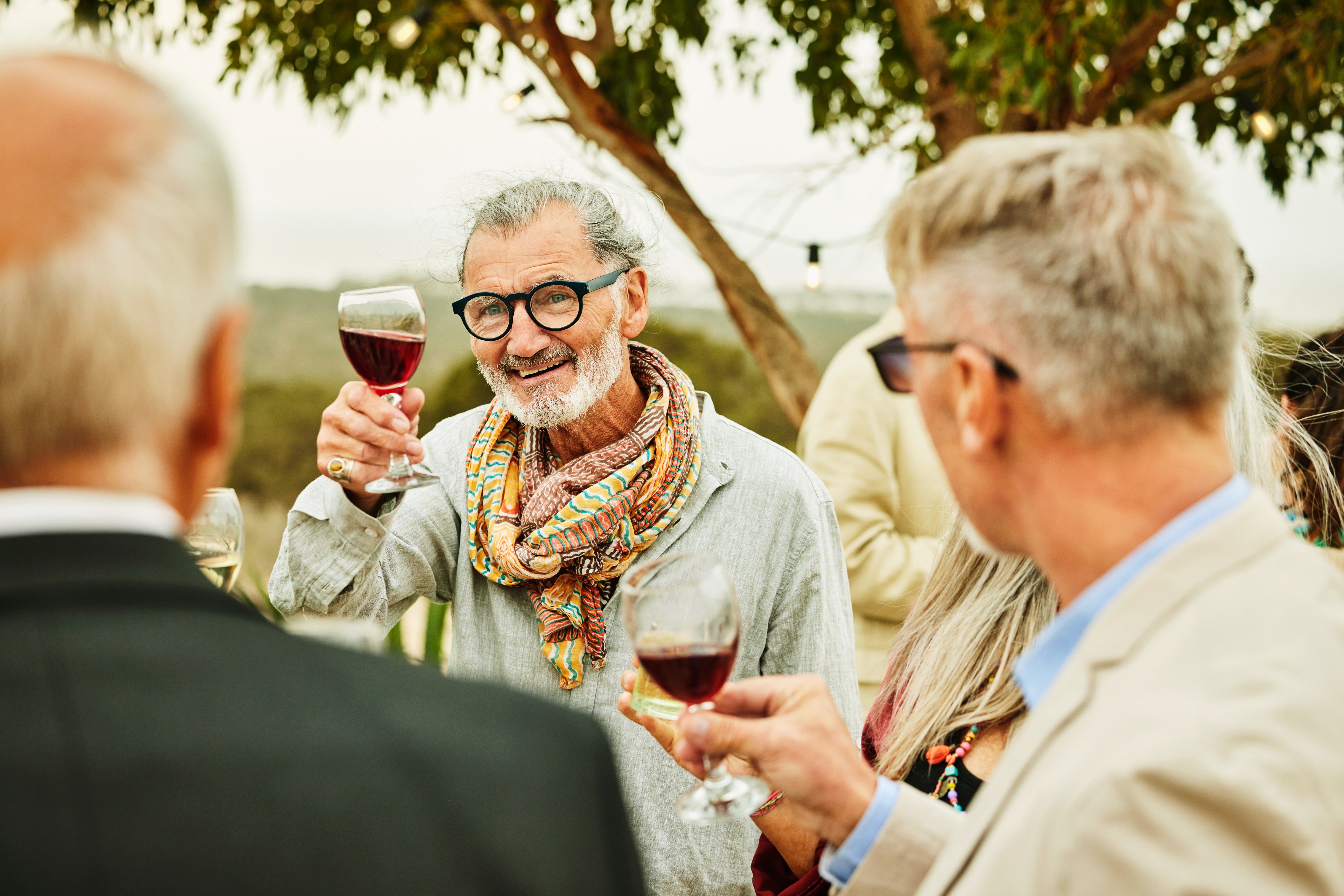 A group of people socialize outdoors, holding wine glasses, with one man in glasses and a patterned scarf raising a toast