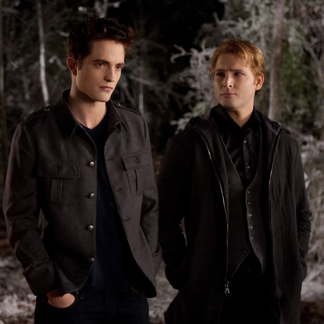 Dos personajes de pie en un bosque nevado, uno con chaqueta casual y el otro con traje más formal. Ambos miran en la misma dirección