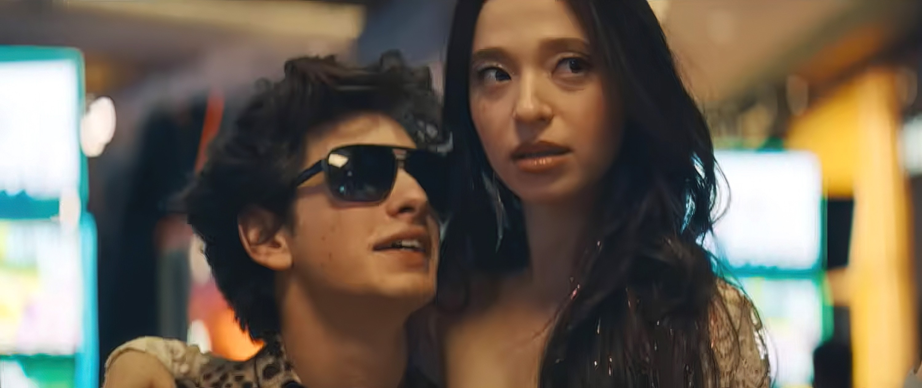 Two people, one wearing sunglasses and a leopard-print shirt, the other in a sparkly outfit, pose together in a lively setting