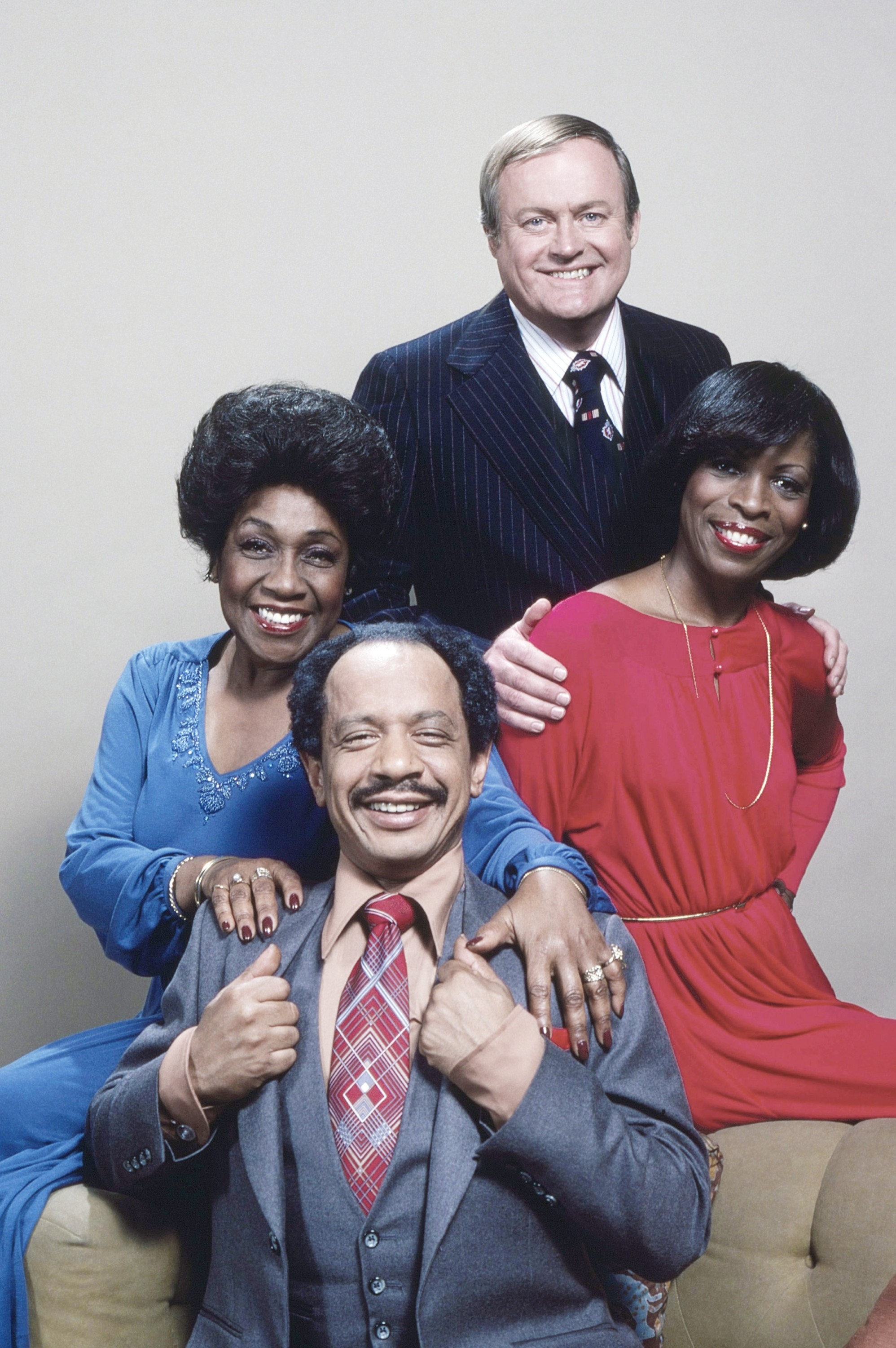 Four people pose together, two seated and two standing. The seated woman wears a blue dress, and the standing woman wears a red dress