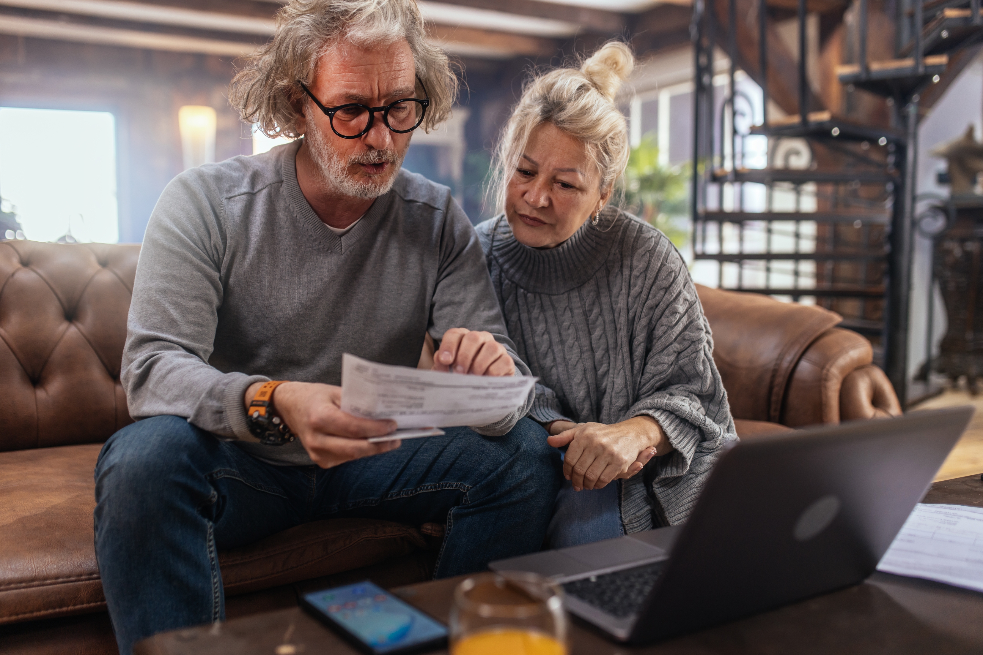 An older couple sitting on a couch reviews paperwork together, with a laptop open on the table in front of them