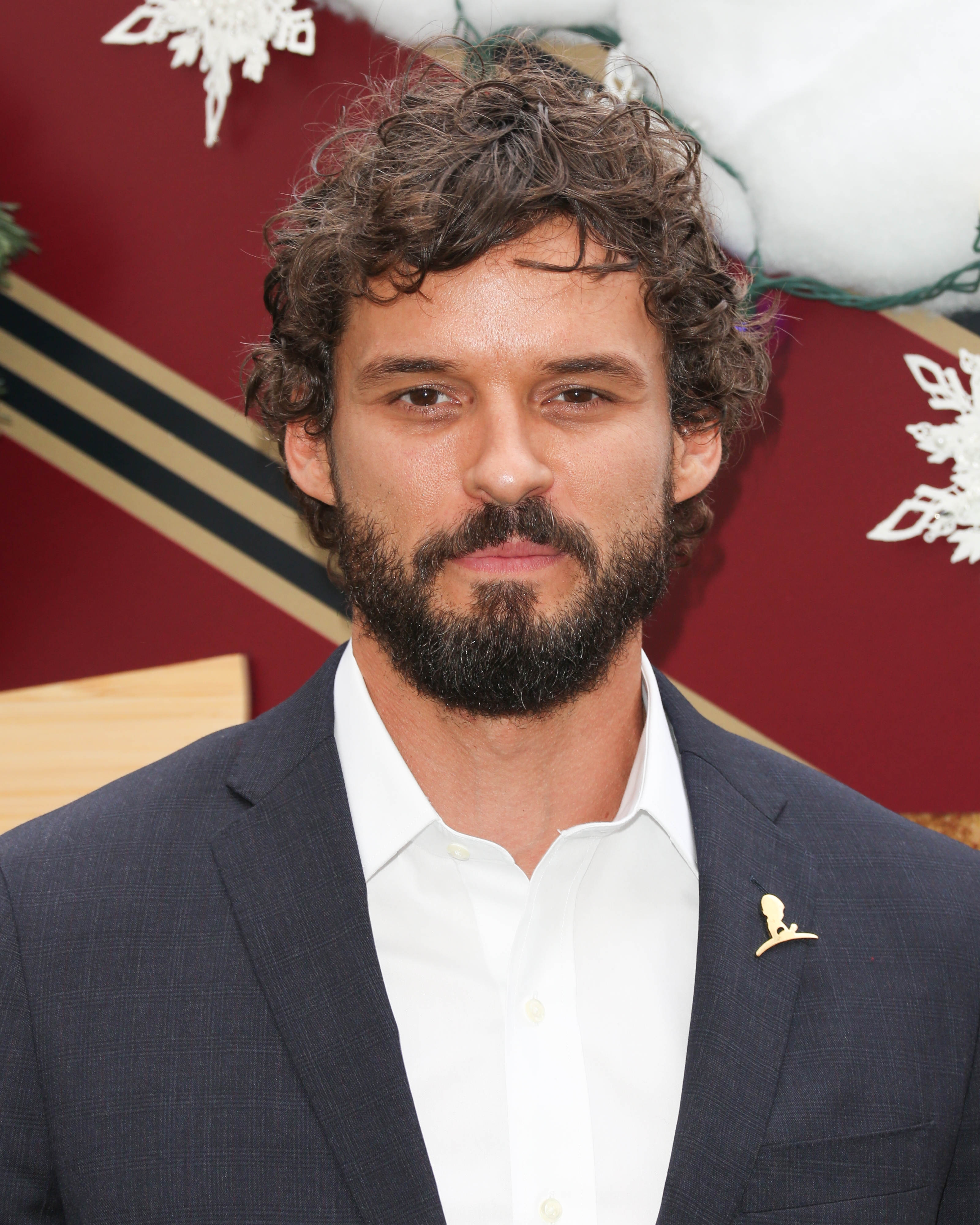 Austin Nichols with curly hair and a beard is wearing a suit jacket and shirt. Snowflake decorations are visible in the background