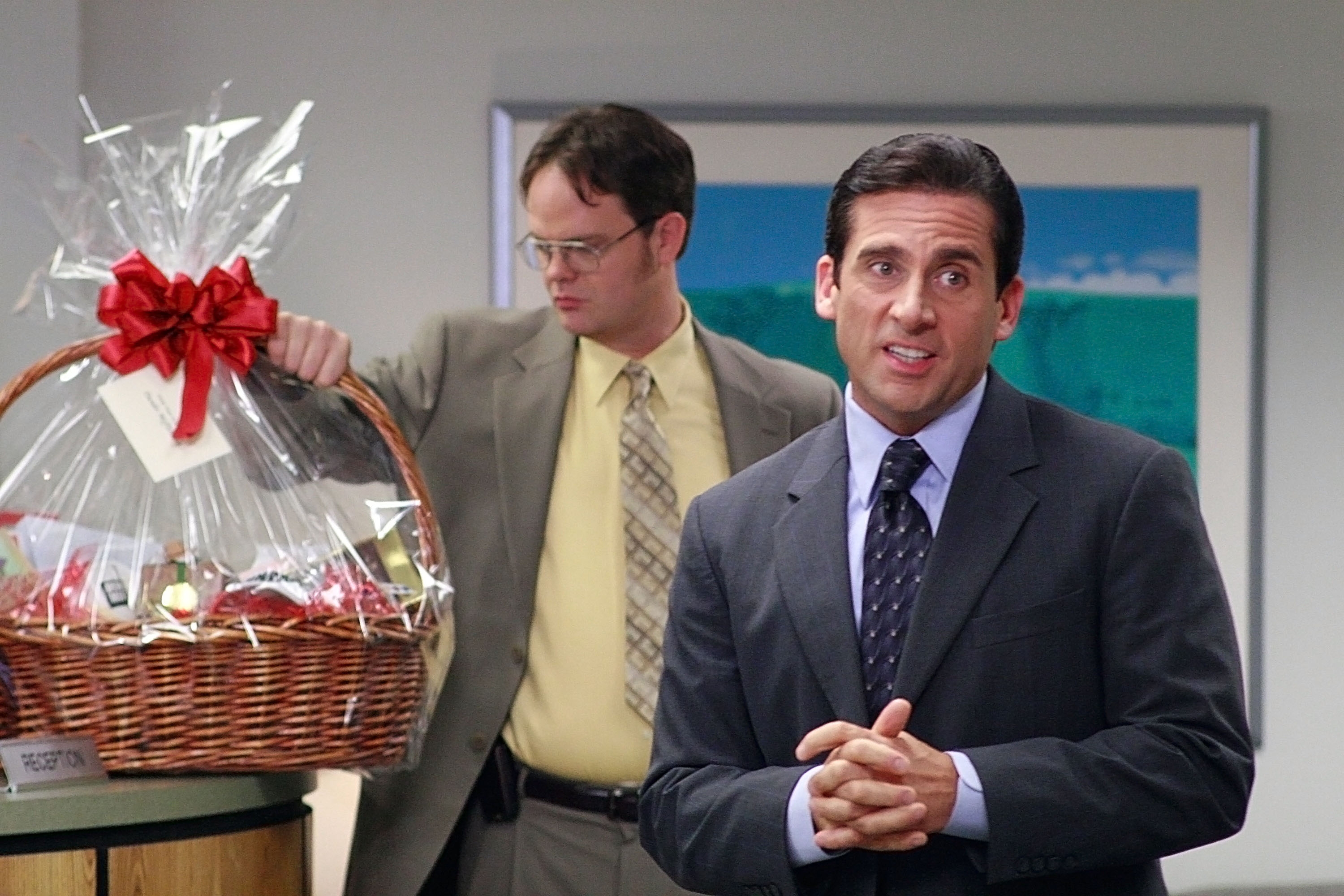 Michael Scott and Dwight in an office setting; one in a suit stands talking, the other examines a gift basket with a bow. Scene from a The Office